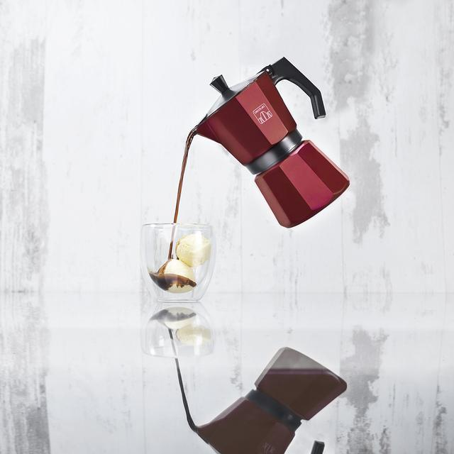 Una cafetera espresso roja vierte café en un vaso con helado, reflejándose en una superficie brillante. Fondo de madera blanca.