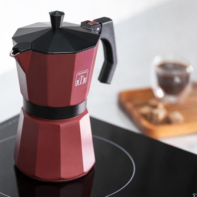 Una cafetera moka roja y negra sobre una vitrocerámica negra. En el fondo, una taza de café y una bandeja de madera.