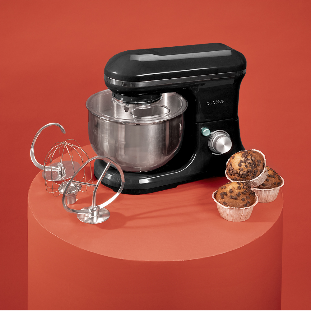 Una batidora negra con accesorios y muffins con chispas de chocolate sobre una superficie roja contra un fondo rojo.