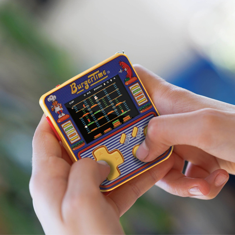 Manos sosteniendo un juego portátil retro de BurgerTime. La pantalla muestra el juego. El dispositivo es amarillo, azul y rojo.