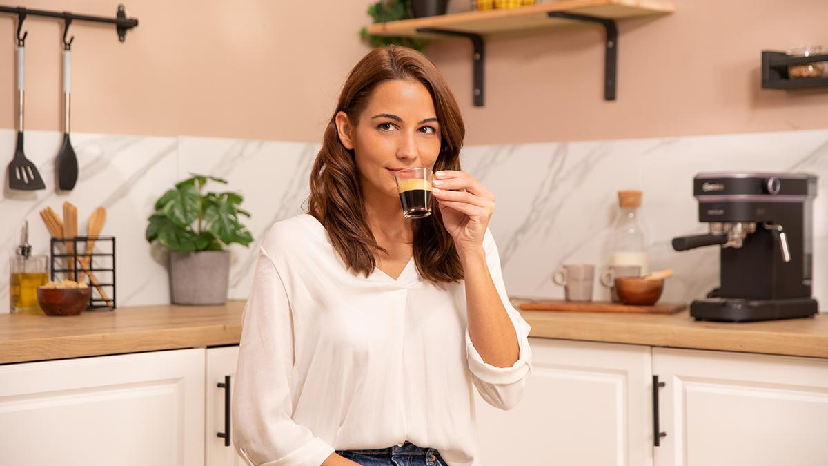 Mujer en camisa blanca sosteniendo un vaso de café espresso. Fondo de cocina con cafetera y utensilios.