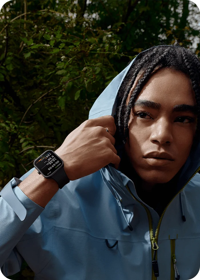 Une personne avec des dreadlocks porte une veste bleue et une montre intelligente. L'arrière-plan montre du feuillage vert.