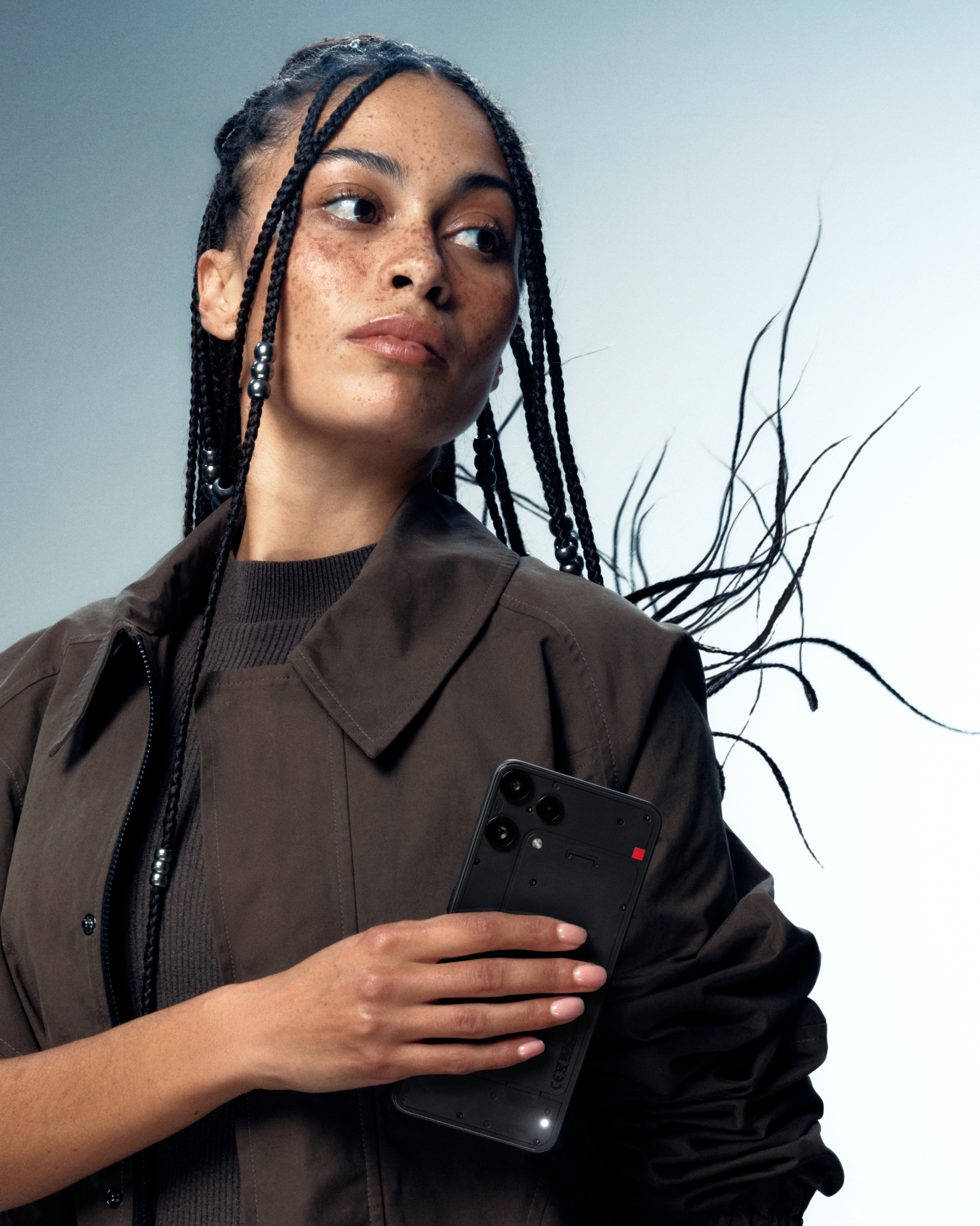Eine Frau hält ein schwarzes Telefon. Sie hat Zöpfe und trägt eine braune Jacke. Der Hintergrund ist hellblau.
