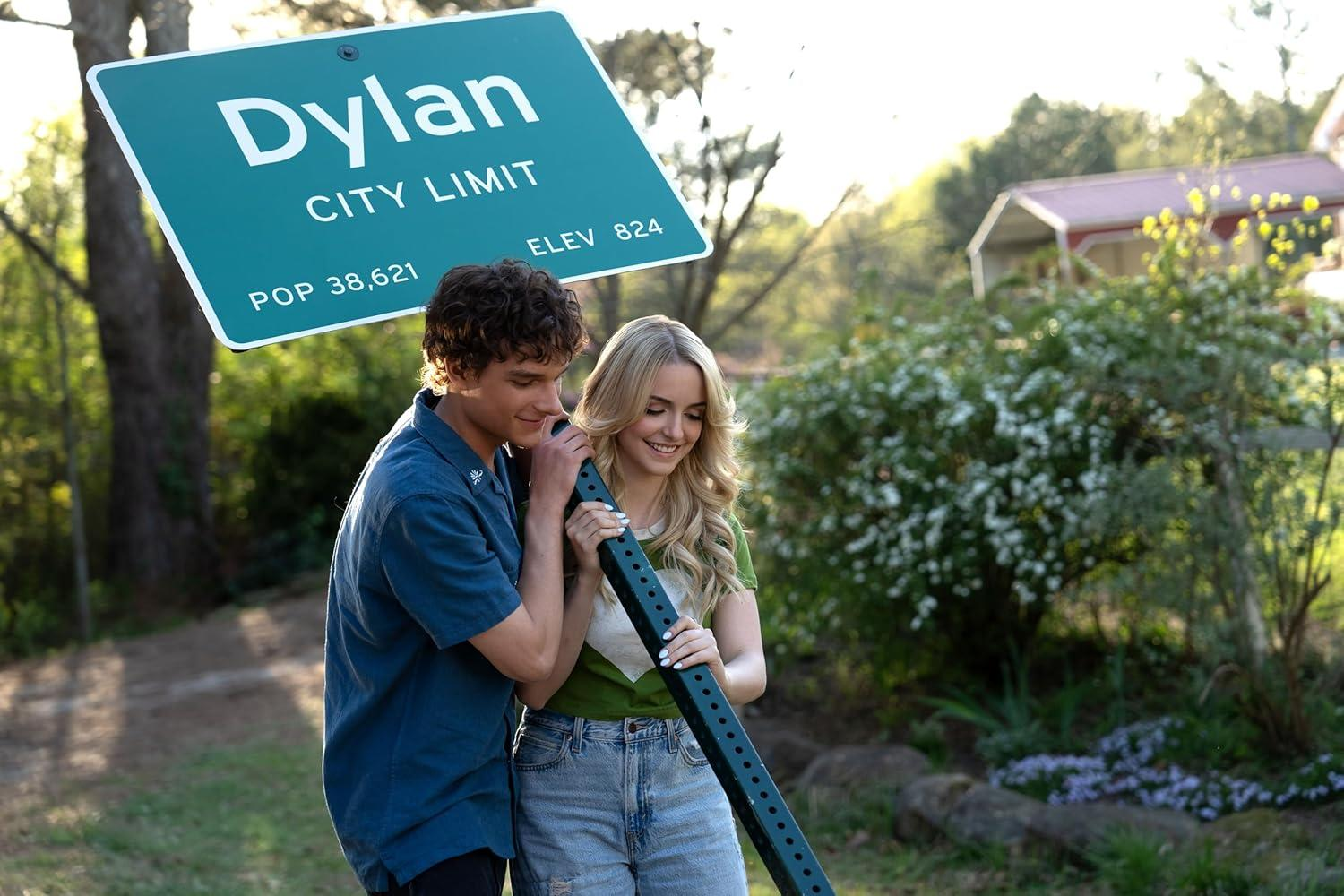 Ein Paar hält ein Ortsschild von Dylan. Das Schild ist über ihnen. Die Frau trägt ein grünes Hemd und Jeans.