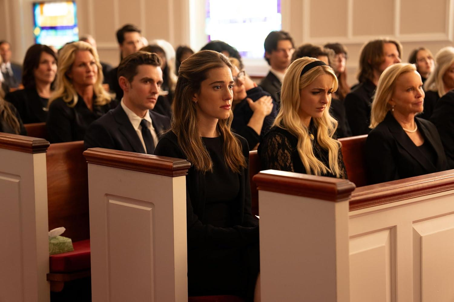 Menschen in einer Kirche. Frauen und Männer in schwarzer Kleidung sitzen in Kirchenbänken. Einige sehen traurig aus. Holzbänke und ein Buntglasfenster sind sichtbar.