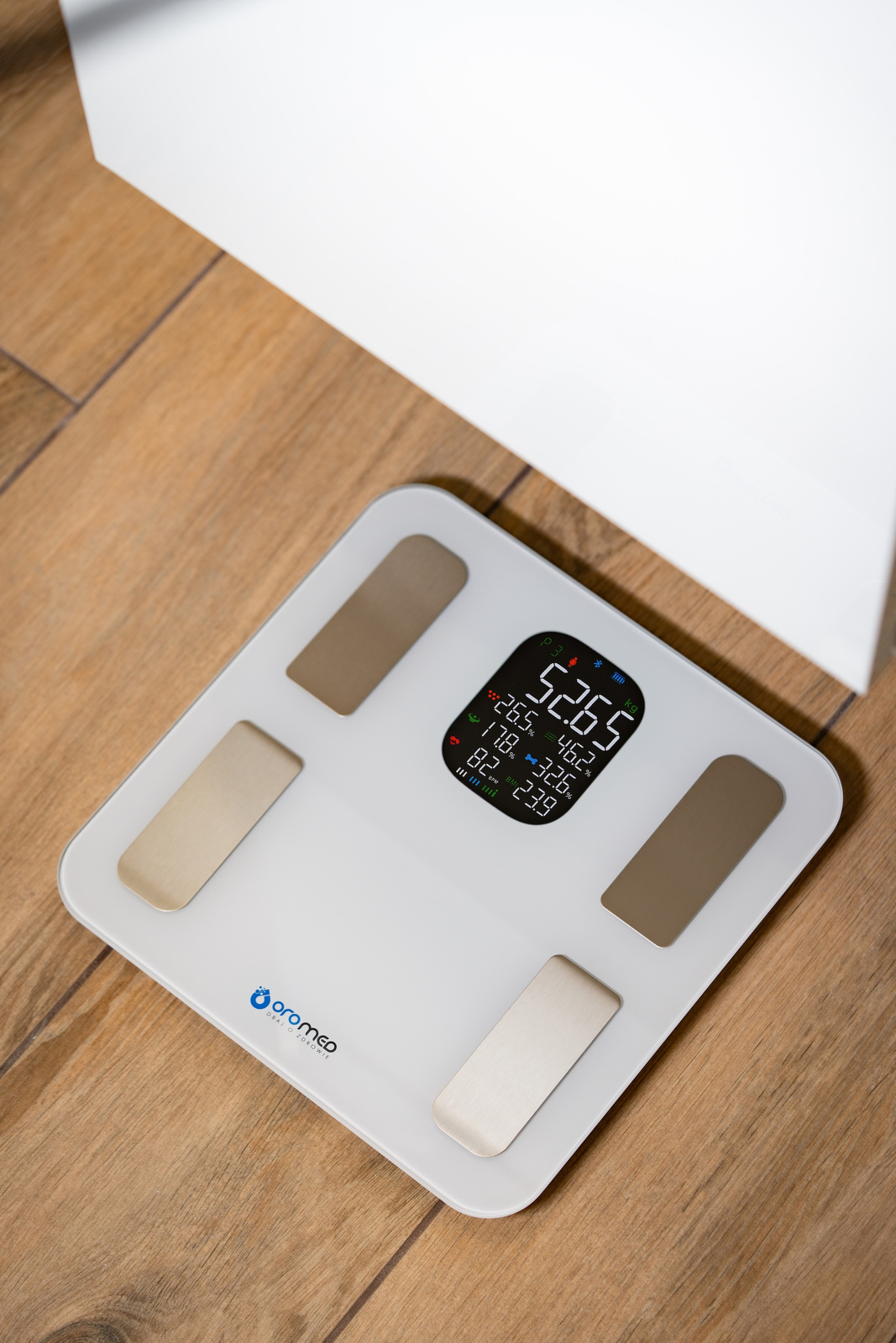 Biała waga cyfrowa na drewnianej podłodze. Pokazuje wagę i skład ciała. Biała kartka na górze.