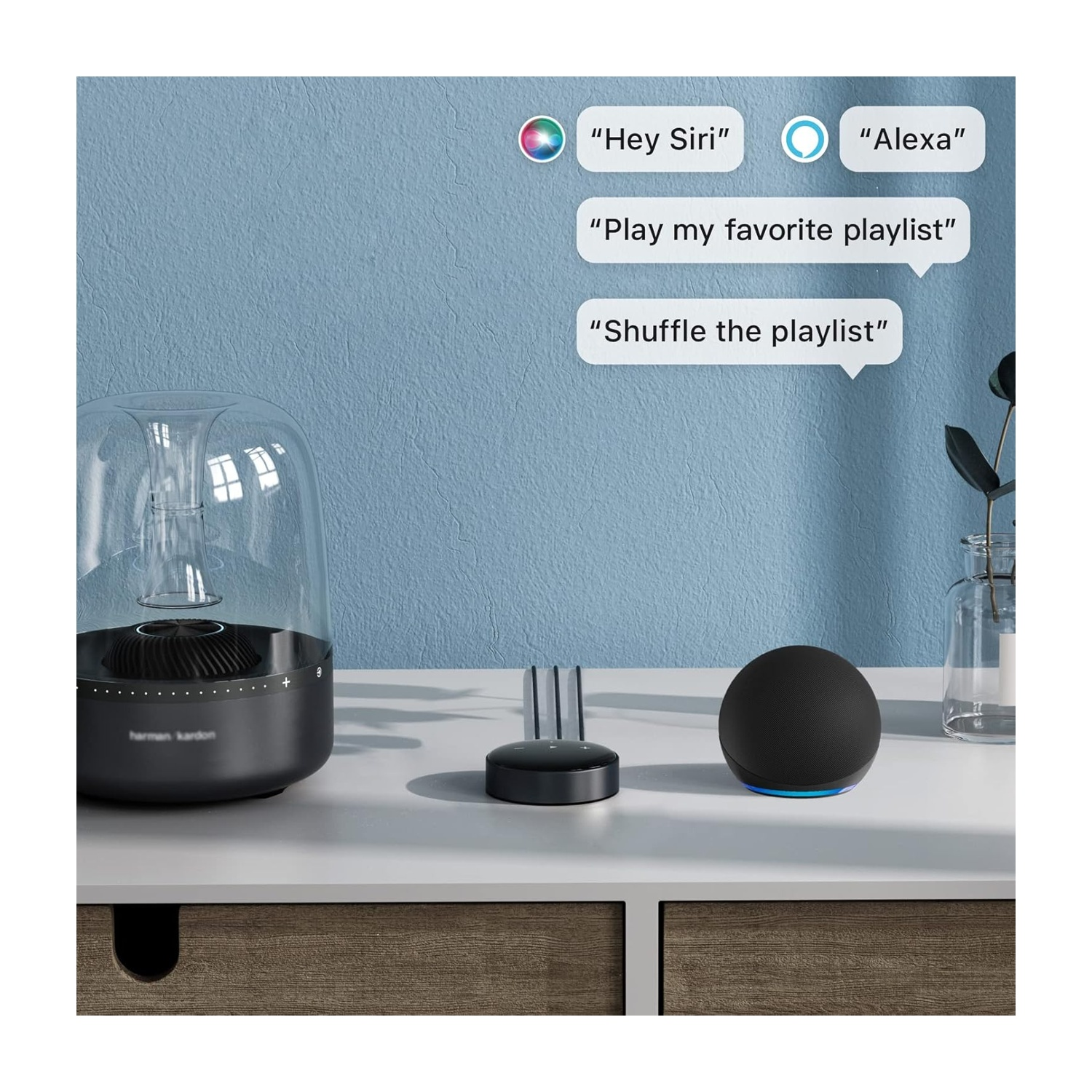 Un altoparlante, un dispositivo Alexa e un piccolo controller nero sono su un tavolo bianco contro un muro blu. Sono presenti fumetti.