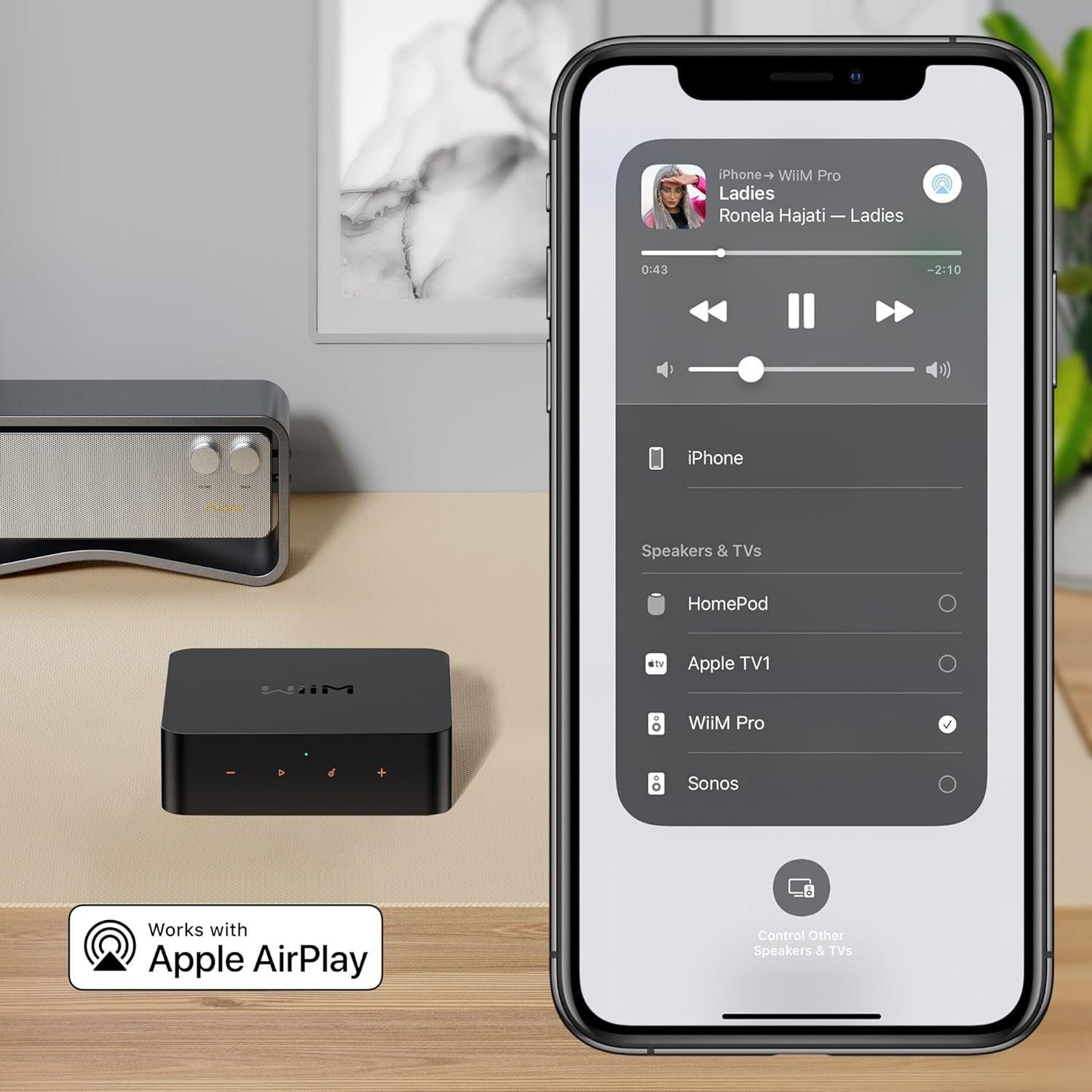 Un iPhone mostra i controlli musicali. Un dispositivo audio nero e un altoparlante sono su un tavolo. Il telefono dice 'Works with Apple AirPlay'.