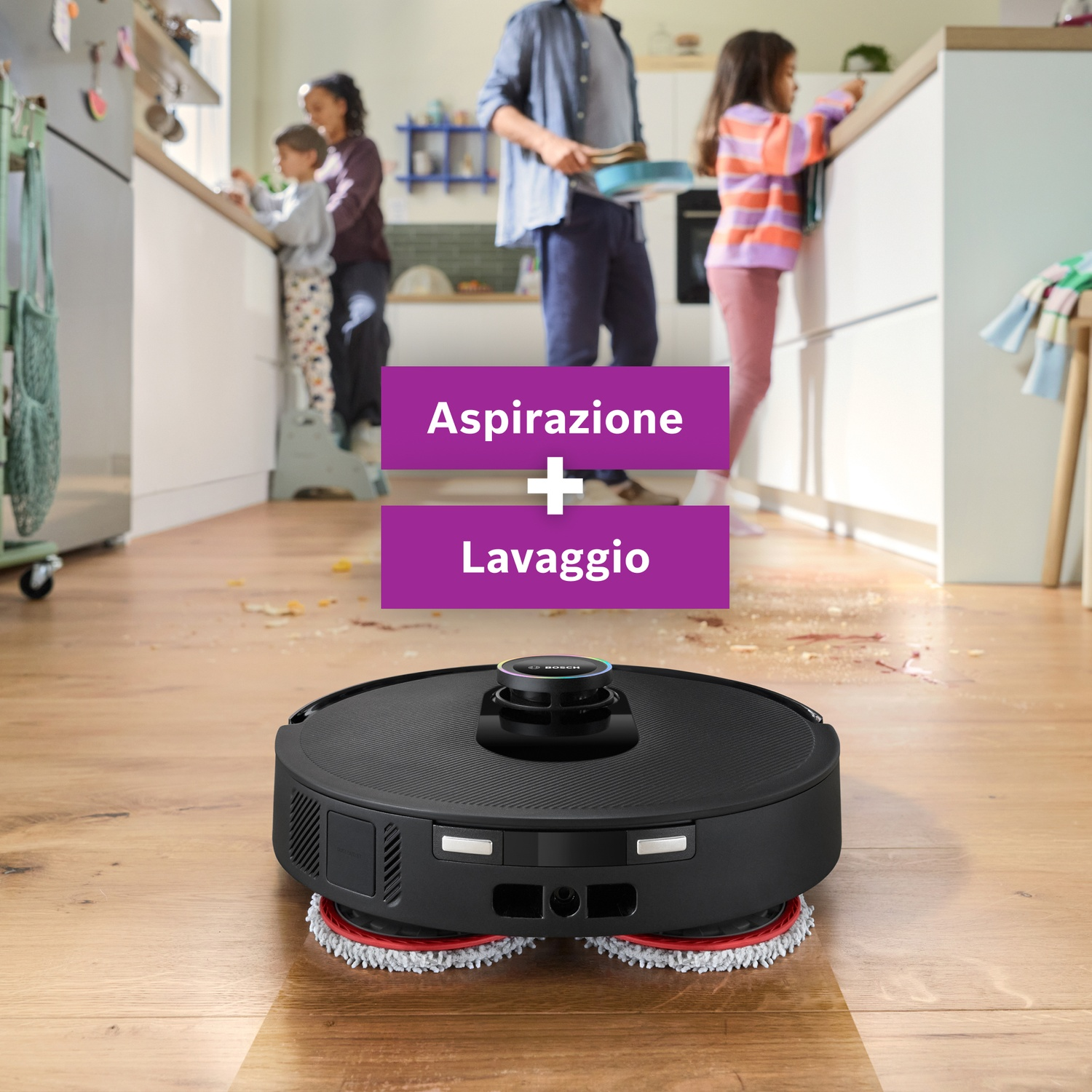 Un aspirapolvere robot nero su un pavimento in legno. Sullo sfondo, una famiglia è in cucina.
