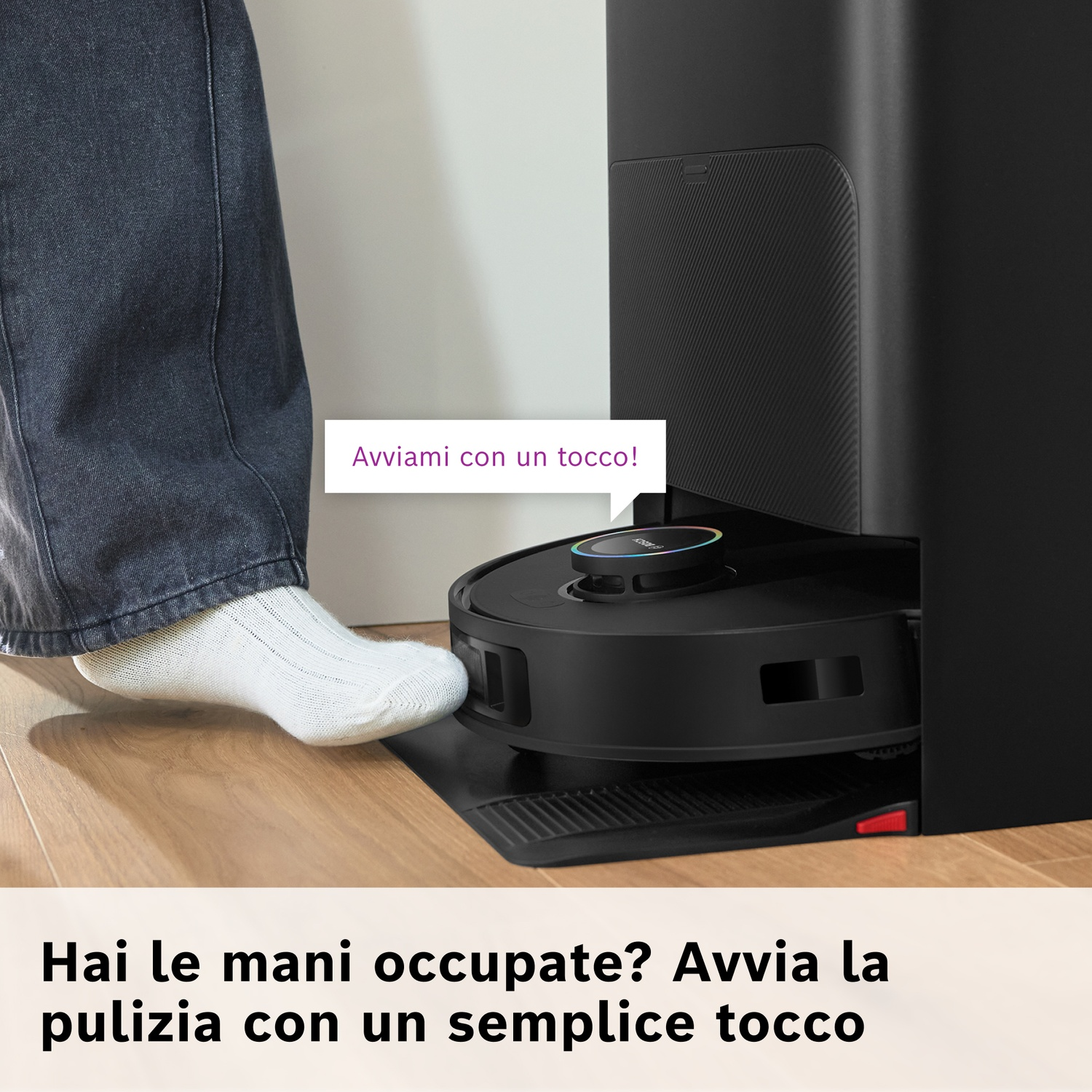 Un piede con un calzino bianco sta per toccare un aspirapolvere robot nero. Dietro c'è una stazione di ricarica nera.