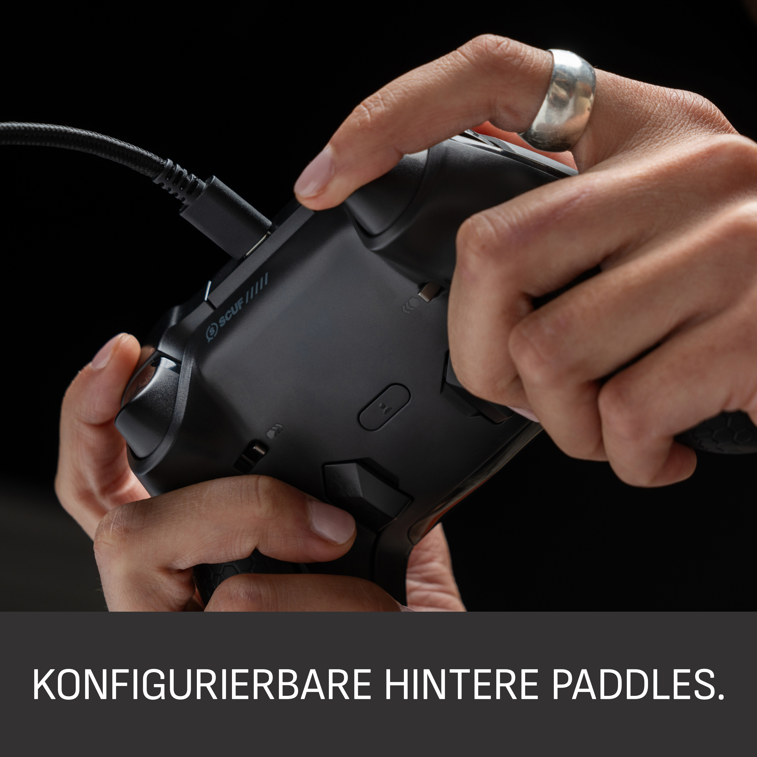 Hände halten einen schwarzen Controller mit eingestecktem Kabel. Ein silberner Ring ist an einem Finger. Schwarzer Hintergrund.