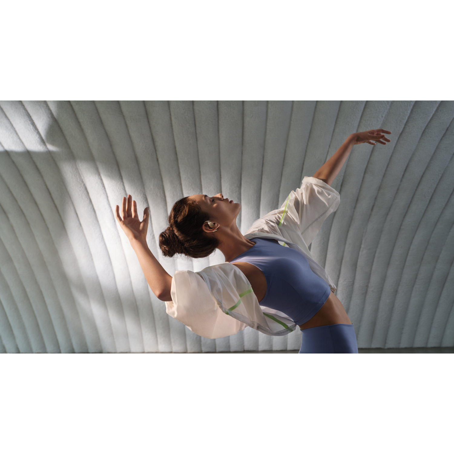 Woman in blue athletic wear dances under a curved textured ceiling. She wears earbuds and a white jacket.