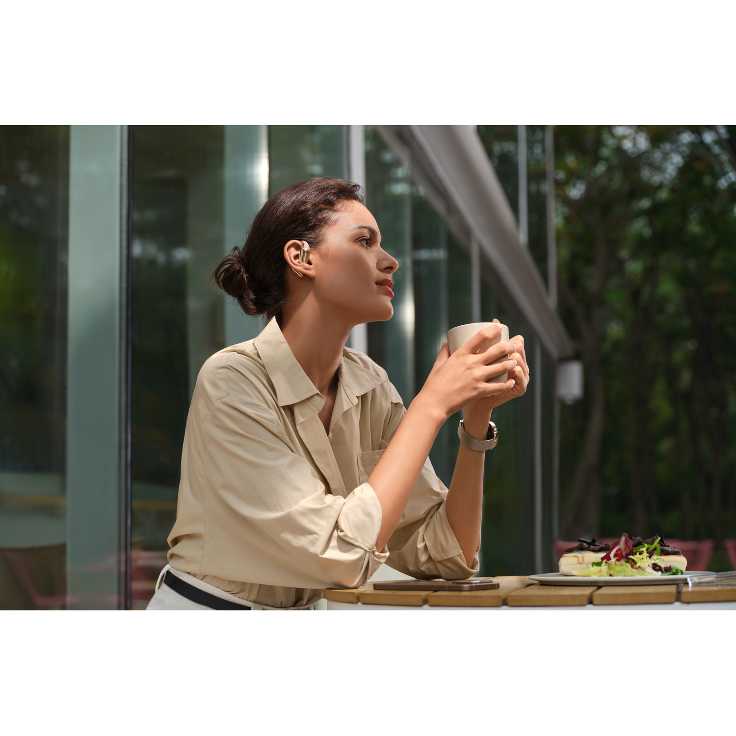 Woman with a bun, holding a cup. Wearing a beige shirt, and a watch. A plate of food is on the table with her.