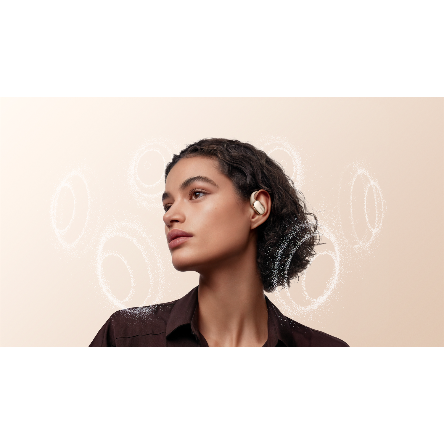 A woman wears a gold earbud. She has dark curly hair and a dark shirt. White swirls are in the background. The skin tone is light.