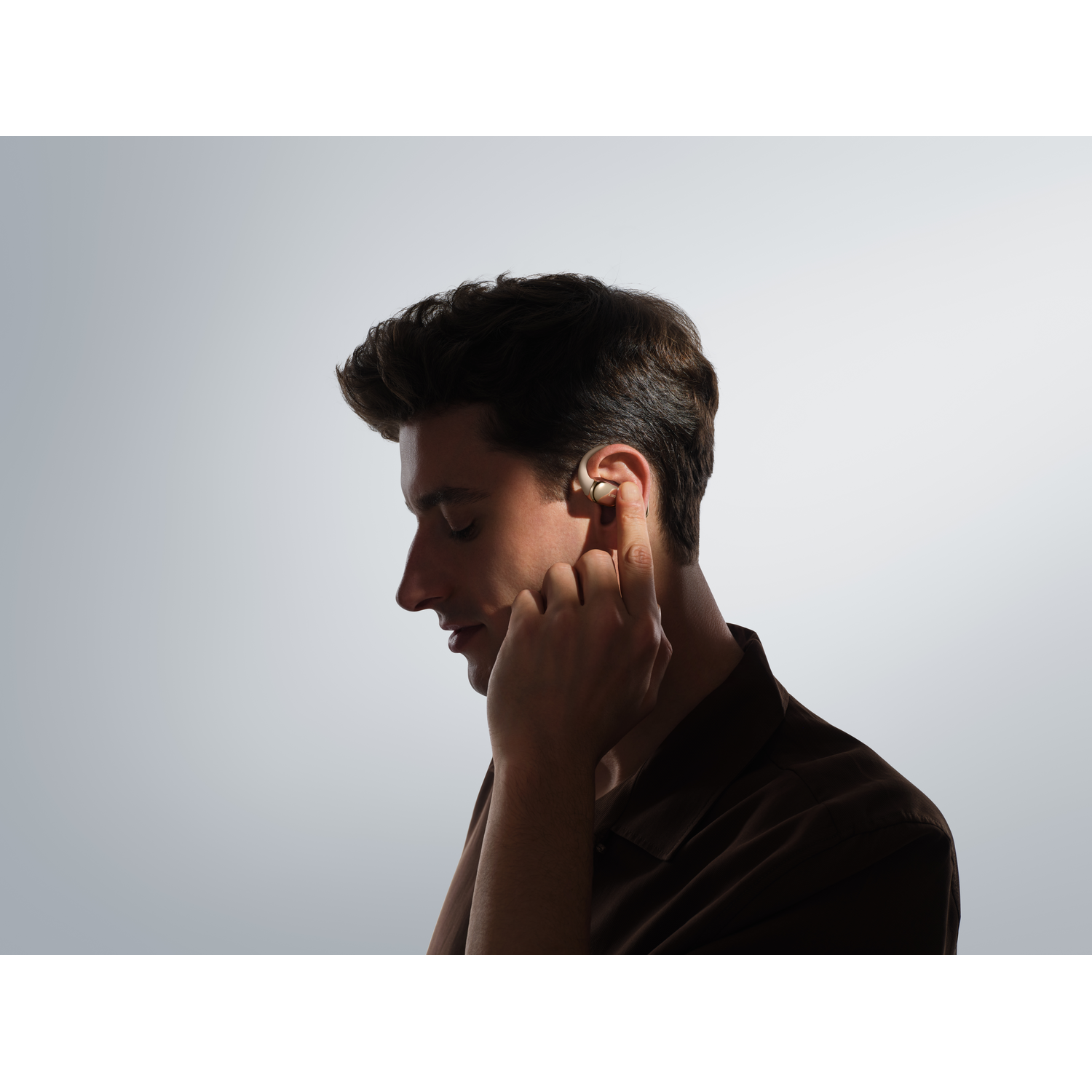 A man in a brown shirt inserts a wireless earbud into his ear, against a gray gradient background.