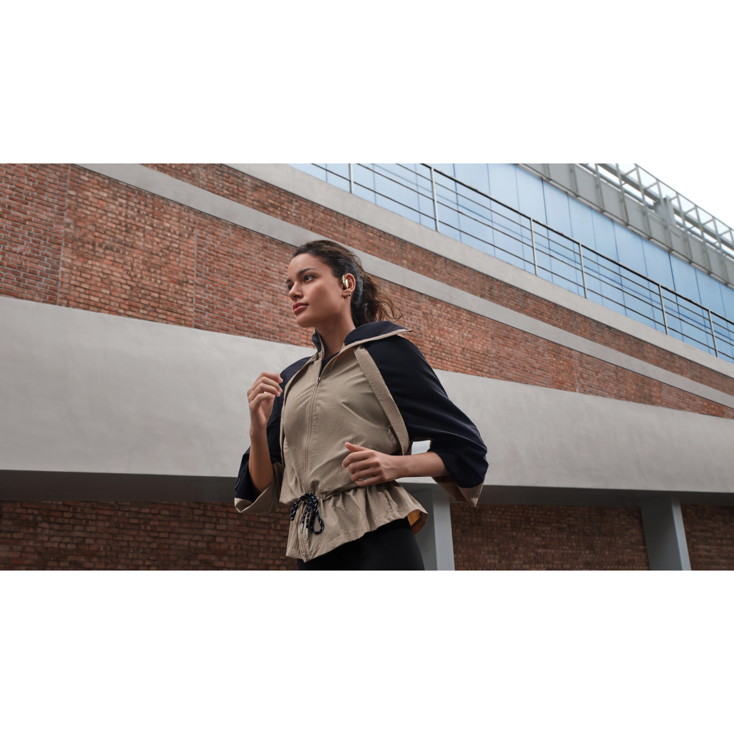 A woman runs wearing earbuds, dressed in a jacket, in front of a brick building and a modern building with glass windows.