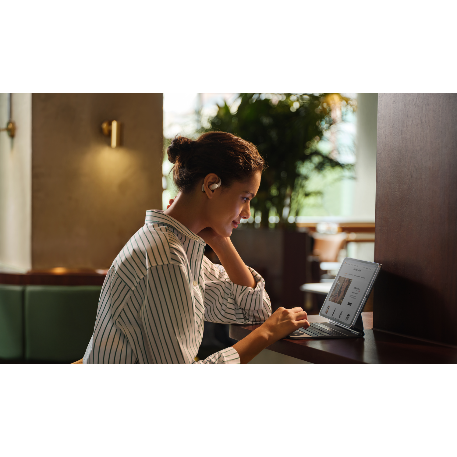 Woman wearing earbuds, working on a laptop in a cafe. She is in a striped shirt. There is a wooden wall and a plant in the back.