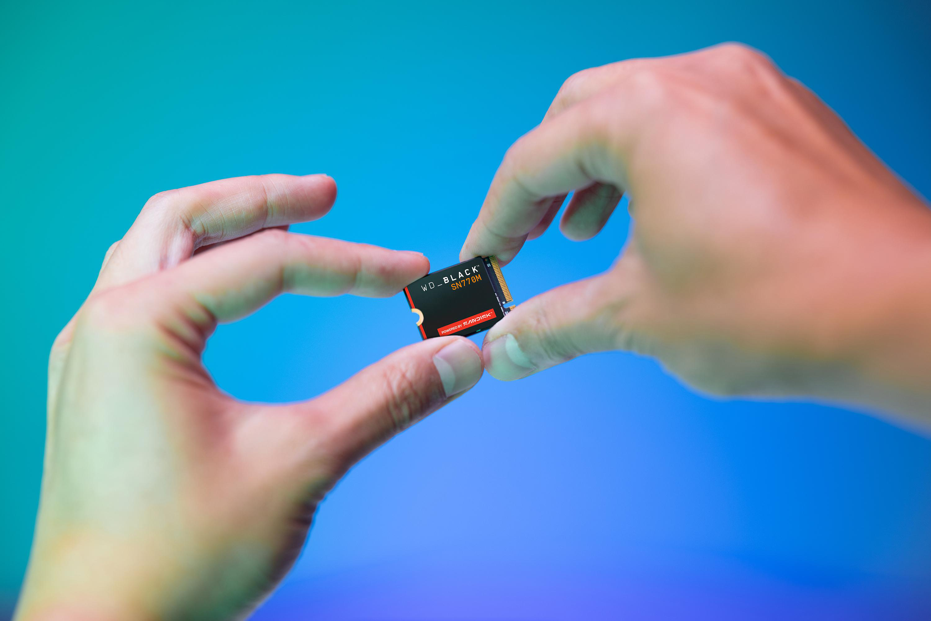 Hände halten eine WD_BLACK SN770M SSD vor einem blau verlaufenden Hintergrund. Die SSD ist schwarz mit roten Akzenten.