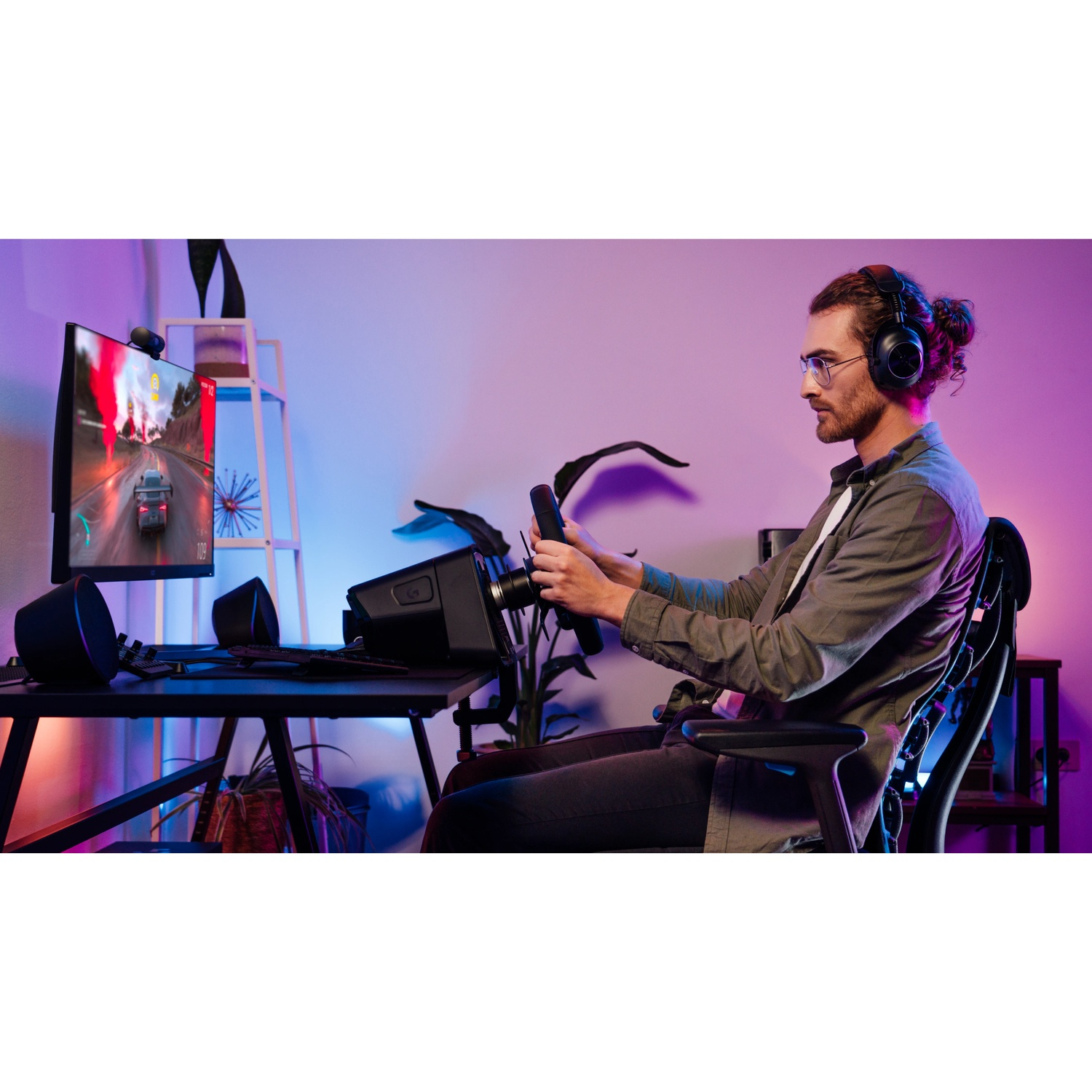 Un uomo gioca a un gioco di corse con un volante e delle cuffie. Lo schermo di un computer mostra una corsa automobilistica. La stanza ha luci colorate.