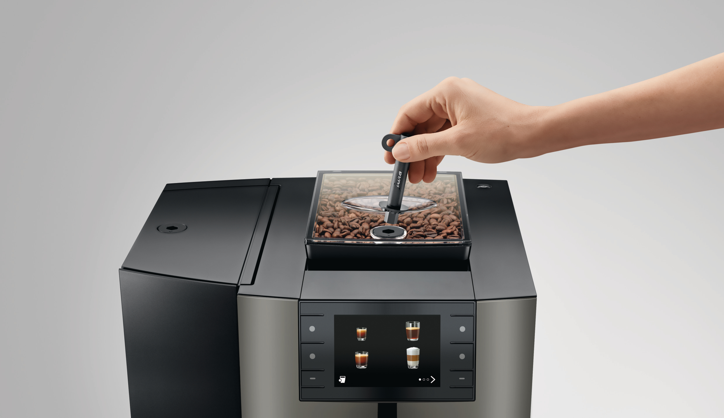 Eine Hand legt einen Bohnenlöffel in den Bohnenbehälter einer Kaffeemaschine. Die Maschine hat einen Touchscreen mit Kaffeeoptionen.