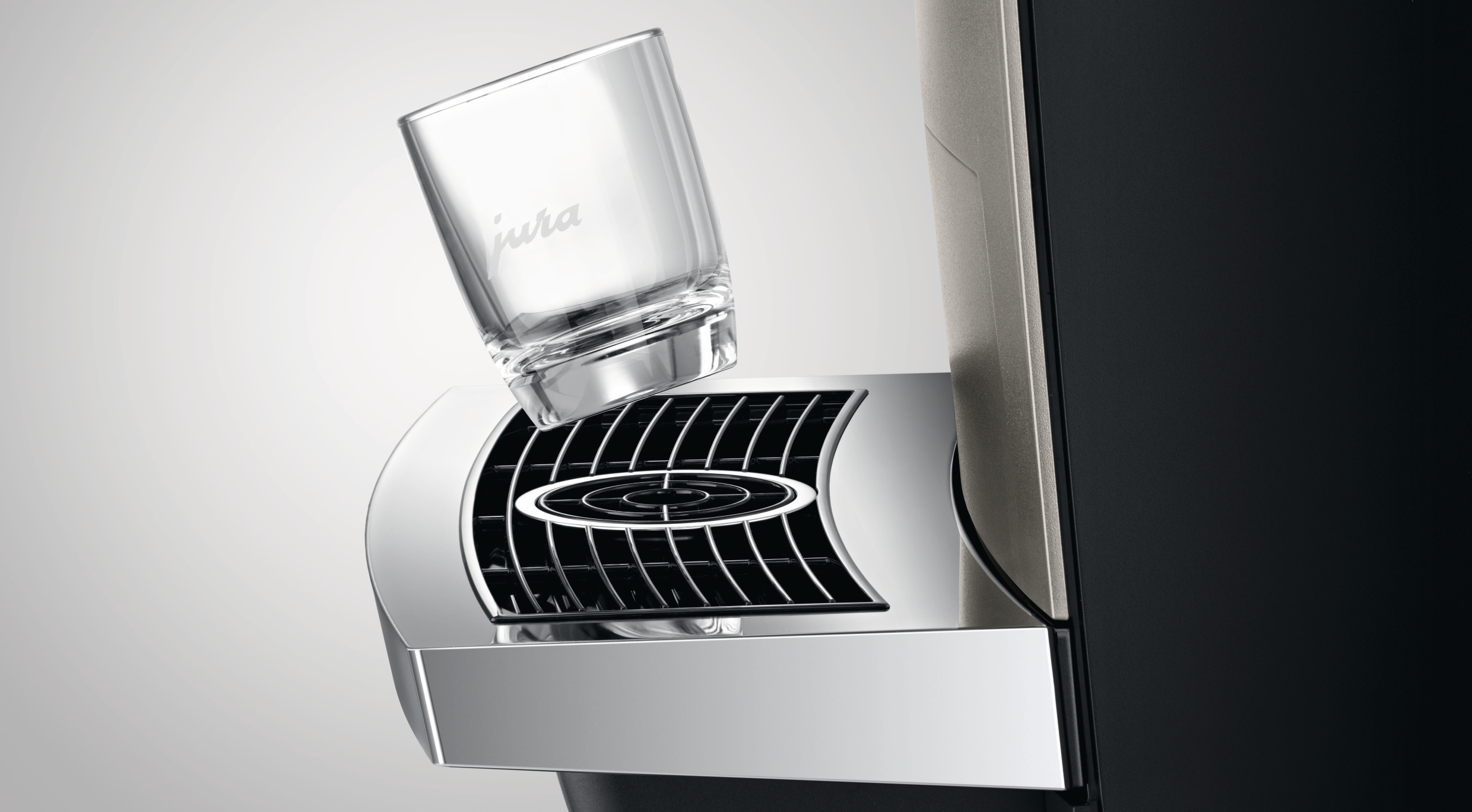Ein klares Glas mit 'jura'-Logo wird über einer Kaffeemaschine platziert. Die Maschine hat einen silbernen Grill und schwarze Teile. Weißer Hintergrund.