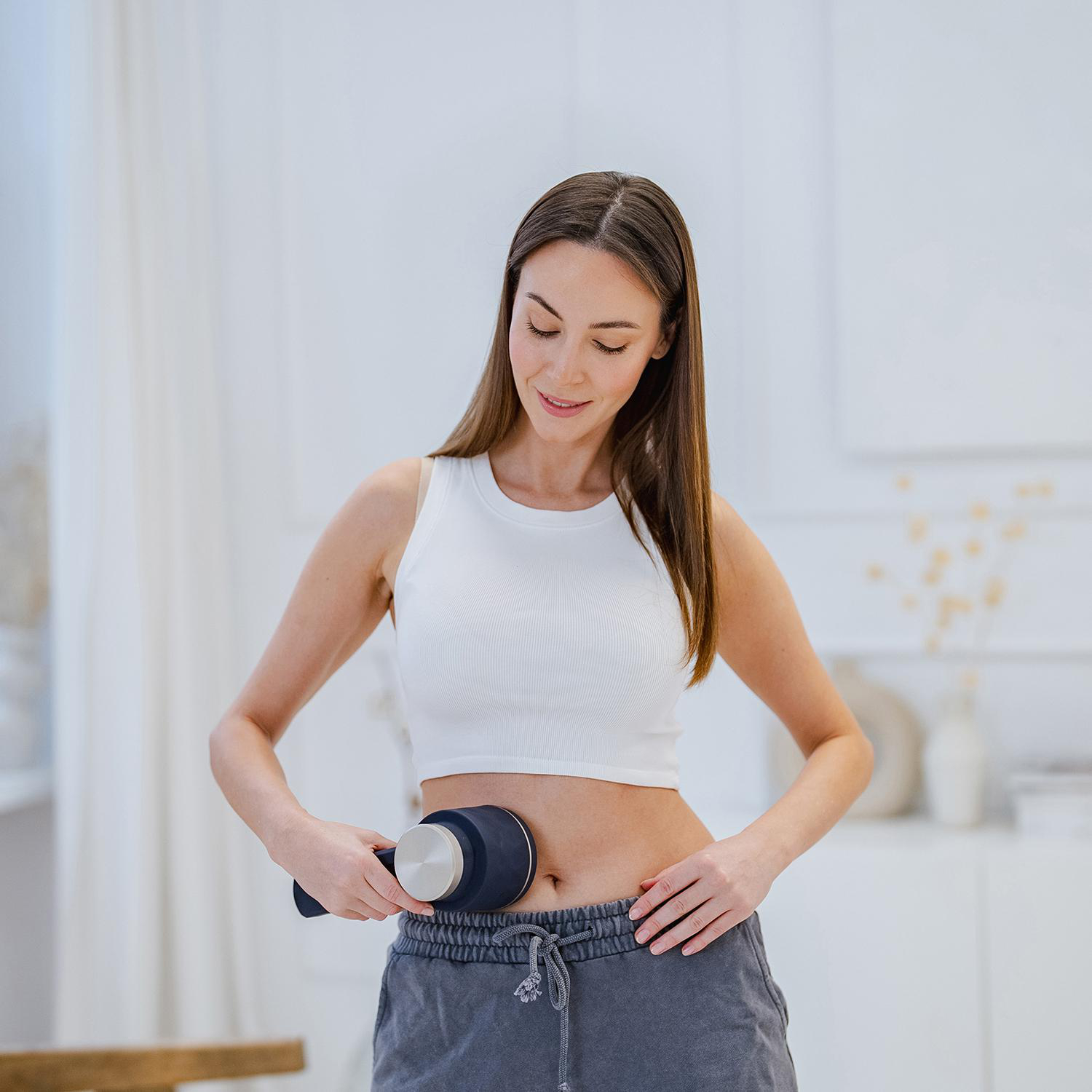 Eine Frau in einem weißen Oberteil und einer grauen Hose hält ein Gerät an ihrem Bauch und lächelt. Der Hintergrund ist ein heller Raum.