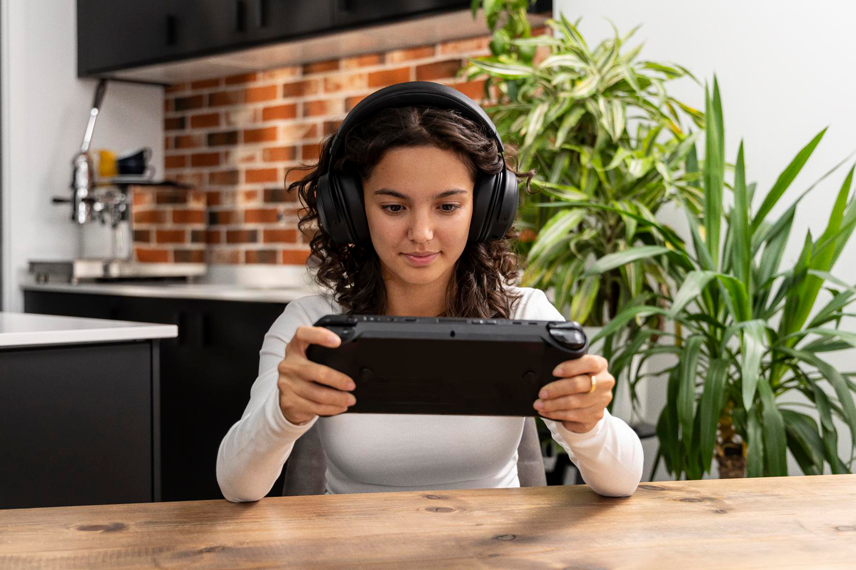 Eine junge Frau mit Kopfhörern spielt ein Handheld-Spiel. Sie sitzt an einem Holztisch vor einer Backsteinmauer.