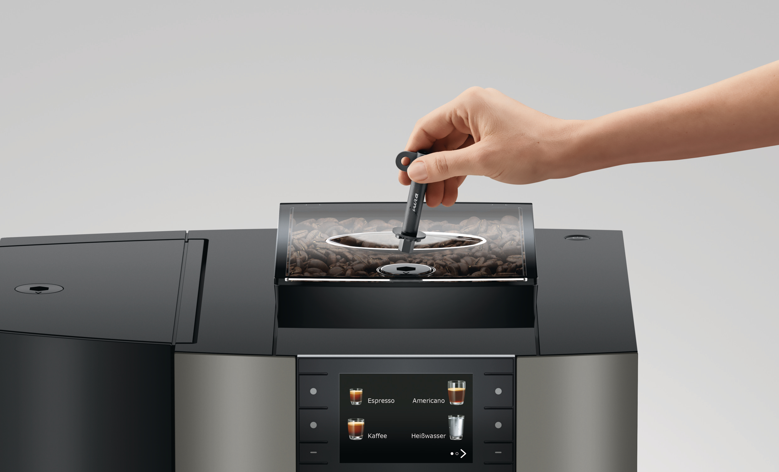 Eine Hand legt ein schwarzes Werkzeug in einen Kaffeemaschinen-Bohnenbehälter. Die Maschine hat einen Touchscreen und Kaffeeoptionen.