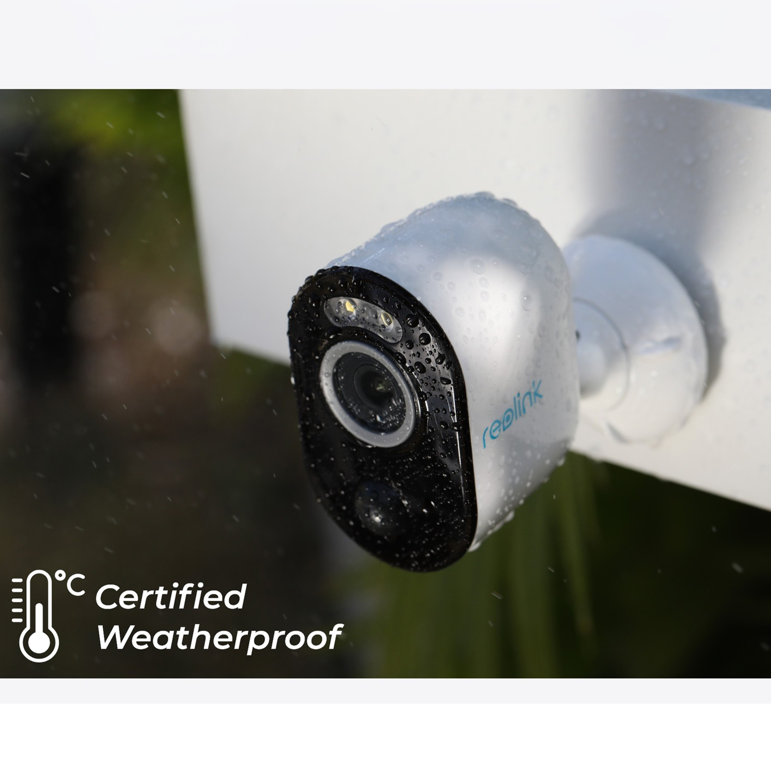 Una telecamera di sicurezza bianca e nera è bagnata da gocce di pioggia. La telecamera è certificata resistente alle intemperie. Fogliame verde sullo sfondo sfocato.
