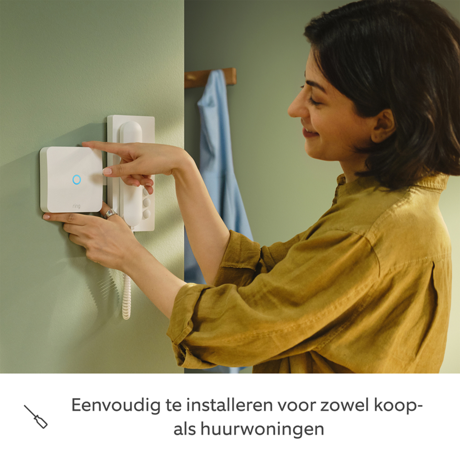 Een vrouw installeert een wit apparaat aan een muur. Ze glimlacht. De muur is groen en een blauw shirt hangt op de achtergrond.