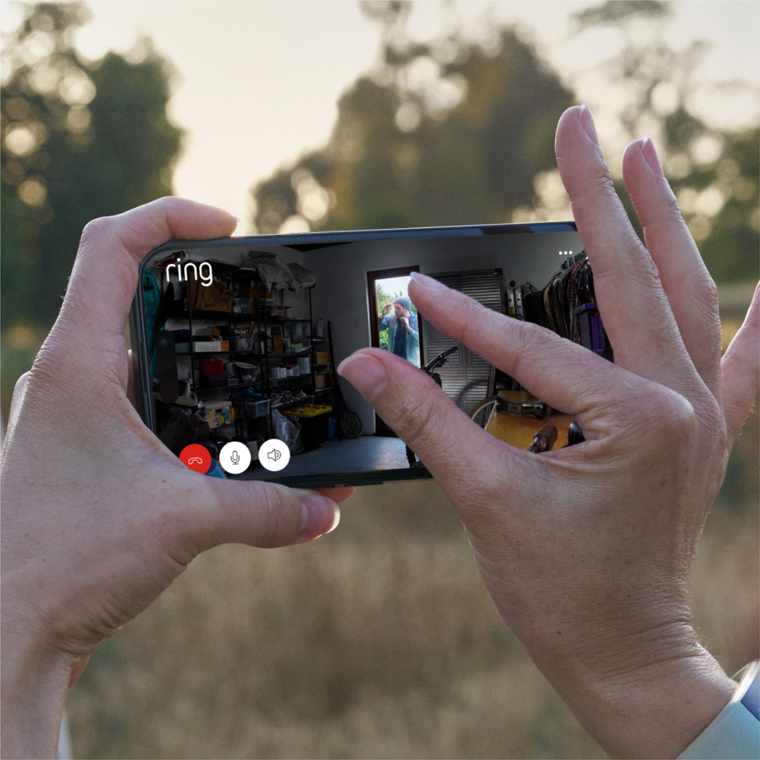Hands holding a phone showing a room via video call. The room has shelves with items, and a person is visible in a doorway.