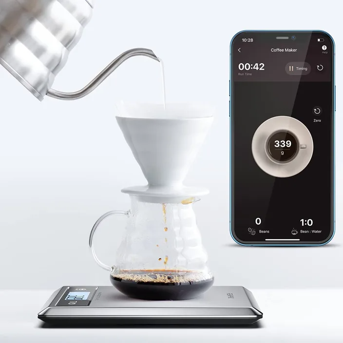 Kaffee wird gebrüht. Wasser gießt aus einem Kessel in einen weißen Filter. Ein Telefon zeigt Brühzeit und Gewicht auf einer Waage.
