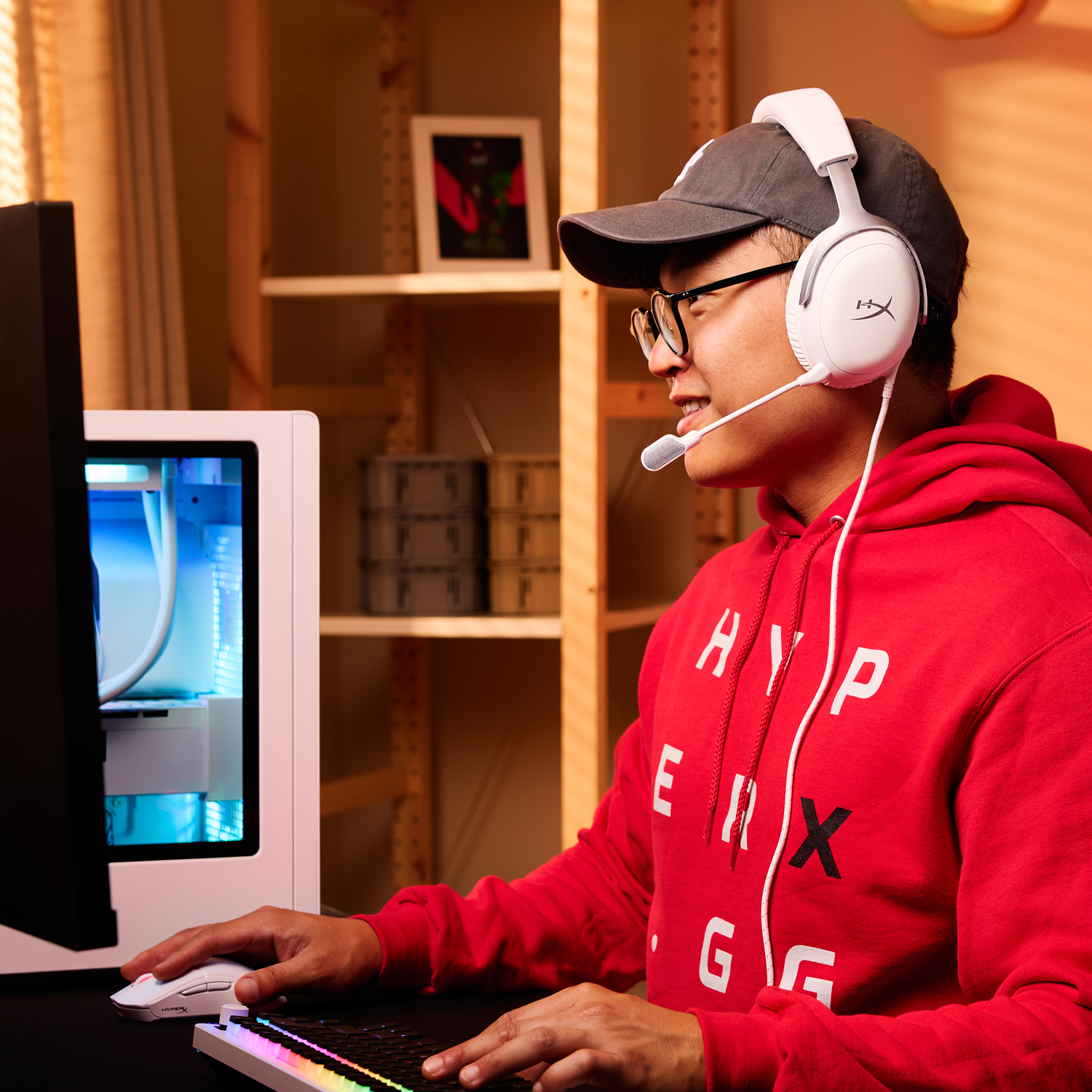 Een man met een headset en bril zit voor een computer. Hij draagt een rode hoodie en een pet. Hij glimlacht.