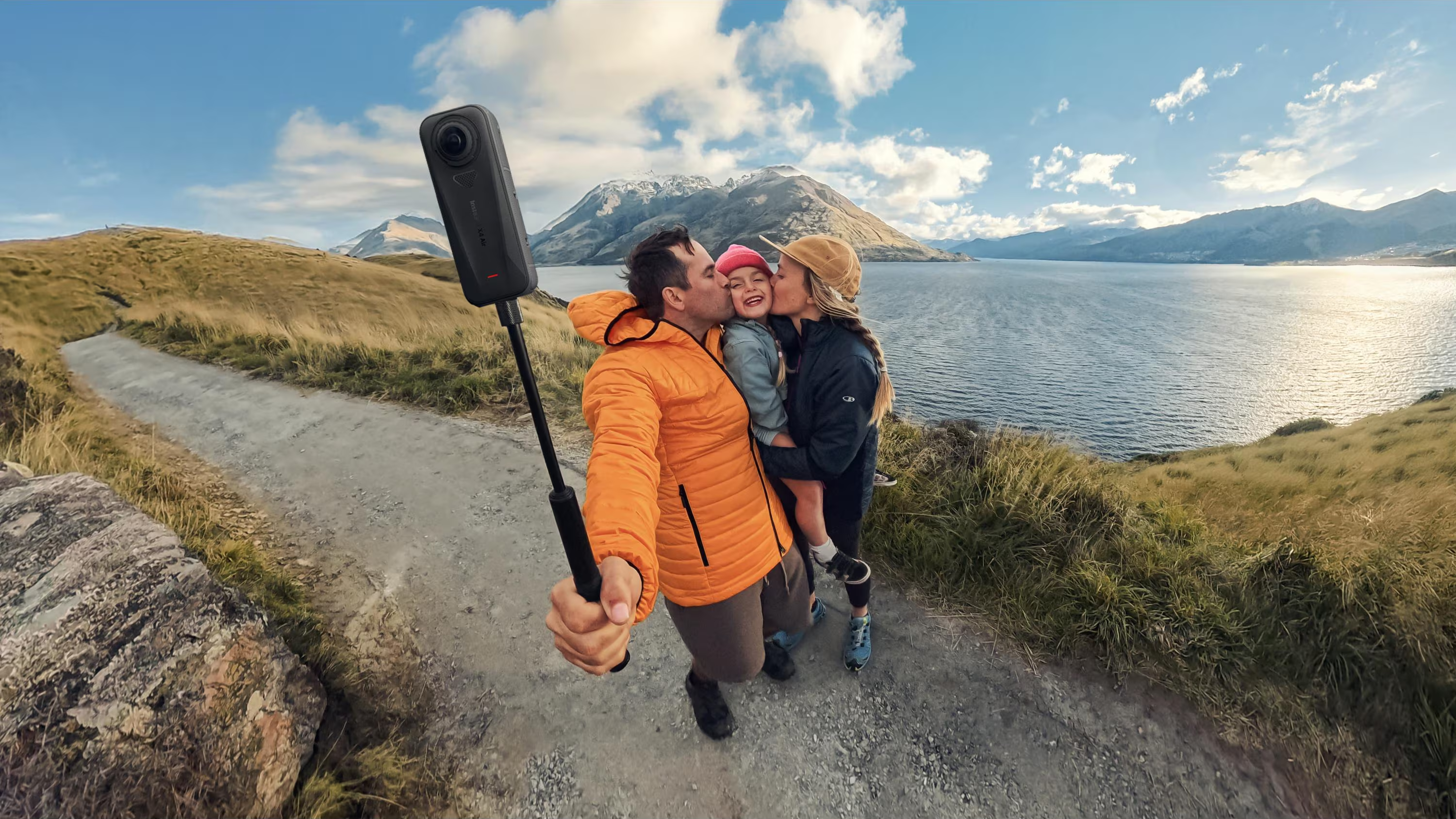 La famiglia scatta una foto con una fotocamera a 360°. Montagne e lago sullo sfondo. Il padre bacia la figlia, la madre la tiene.