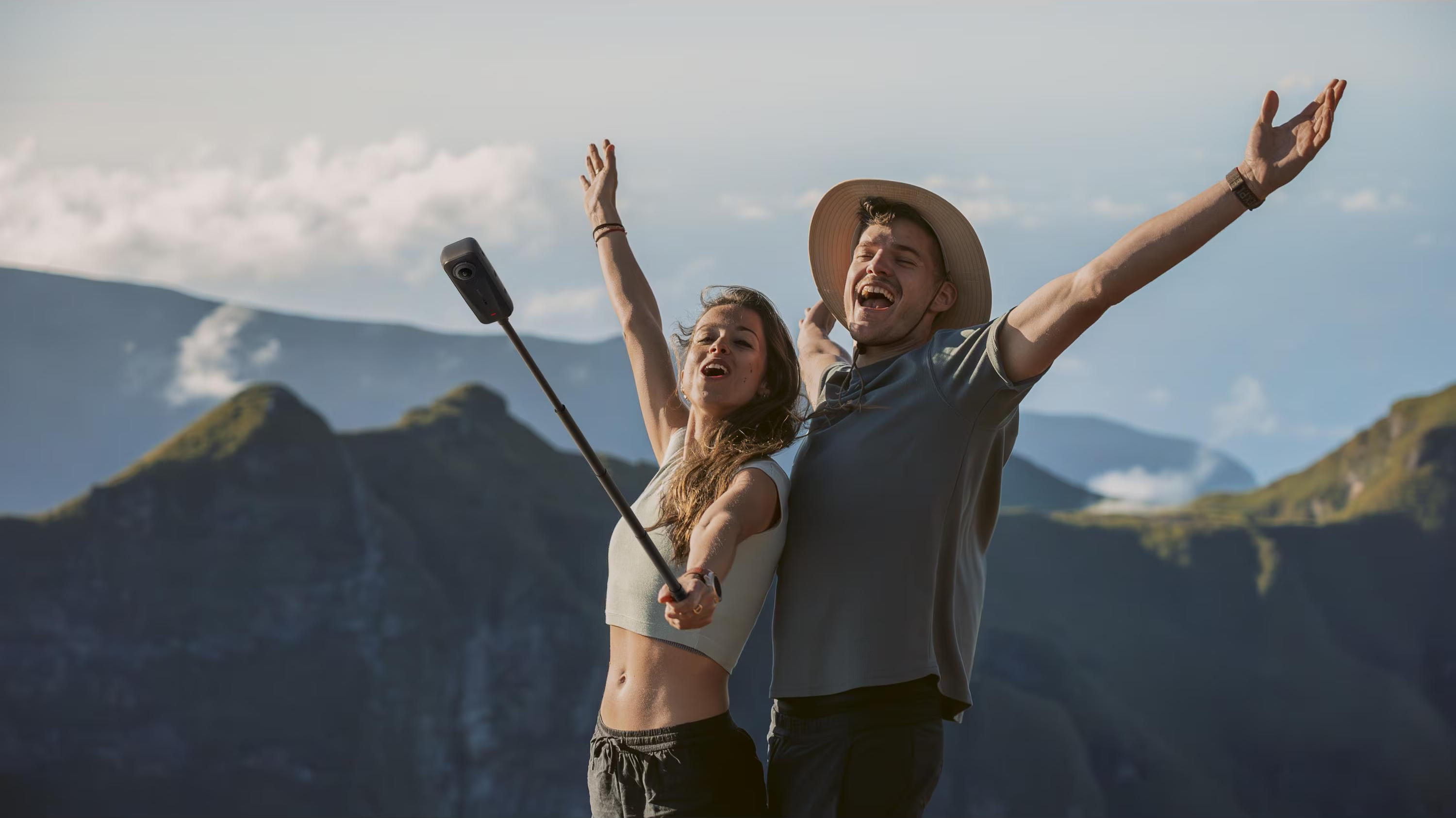 Una donna e un uomo gioiosi, con le braccia alzate, sullo sfondo di montagne. Uno tiene un bastone per selfie.
