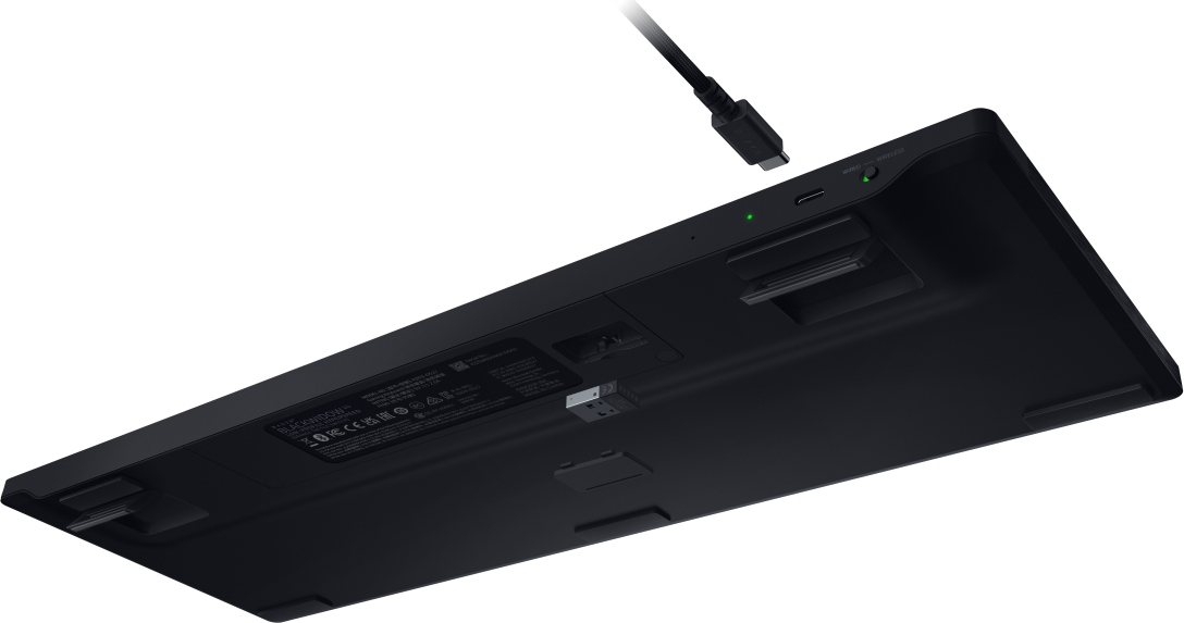 Tastiera nera con cavo USB-C collegato. Luce verde accesa. Vista inferiore con etichette e piedini.