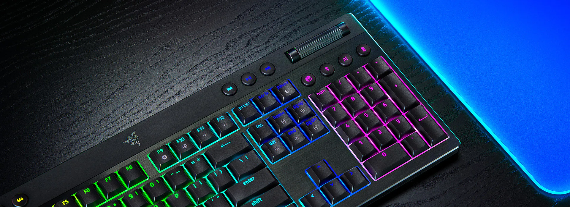 Tastiera Razer nera con tasti retroilluminati colorati su una superficie di legno nero, un tappetino blu a destra.