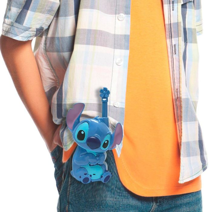 Una persona sostiene un juguete azul de Stitch, con una antena de flor, enganchado al bolsillo de su camisa. Viste una camisa a cuadros y una camiseta naranja.