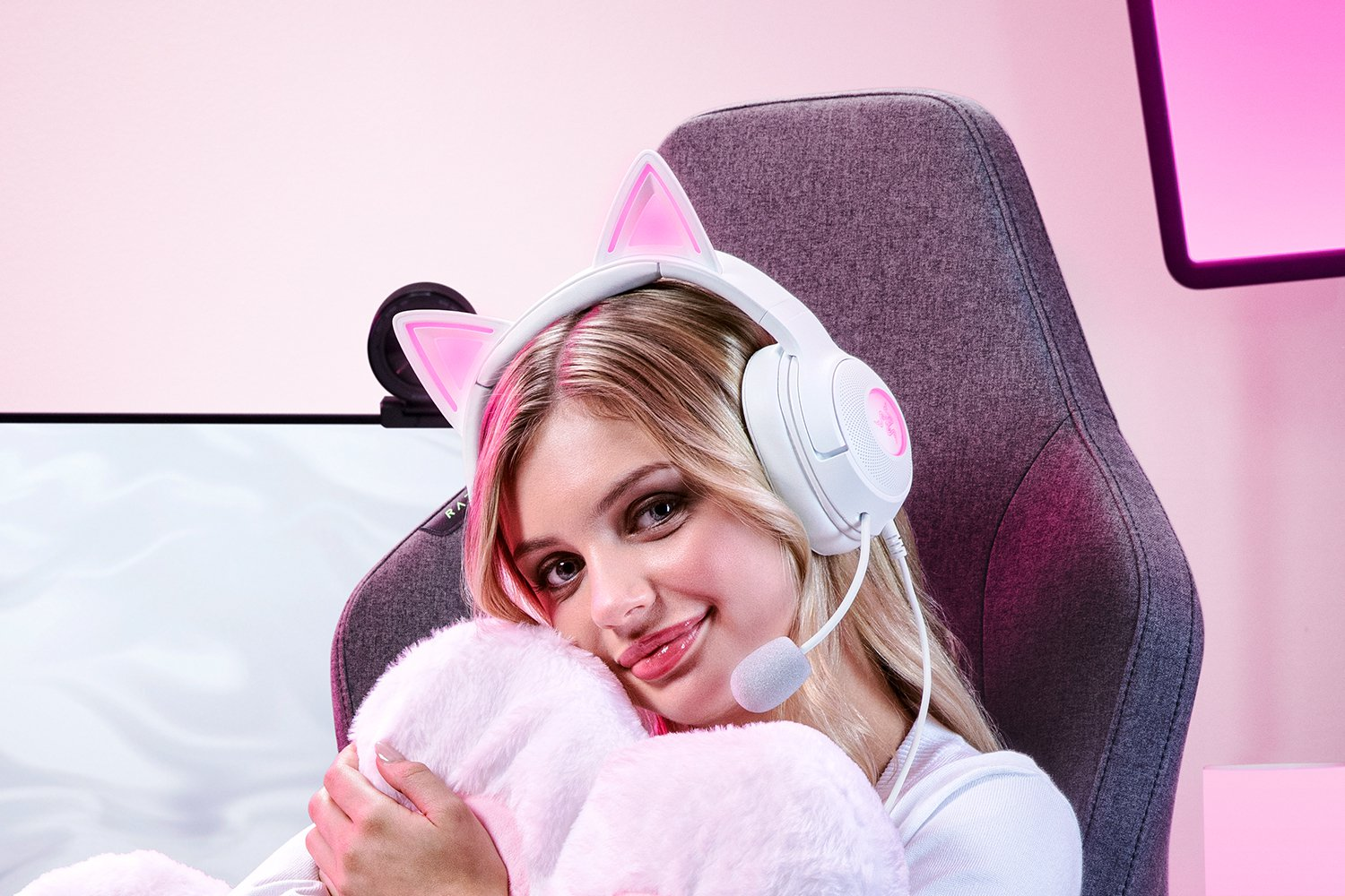Joven con auriculares blancos con orejas de gato, sosteniendo un peluche. Sonríe sentada en una silla gris.