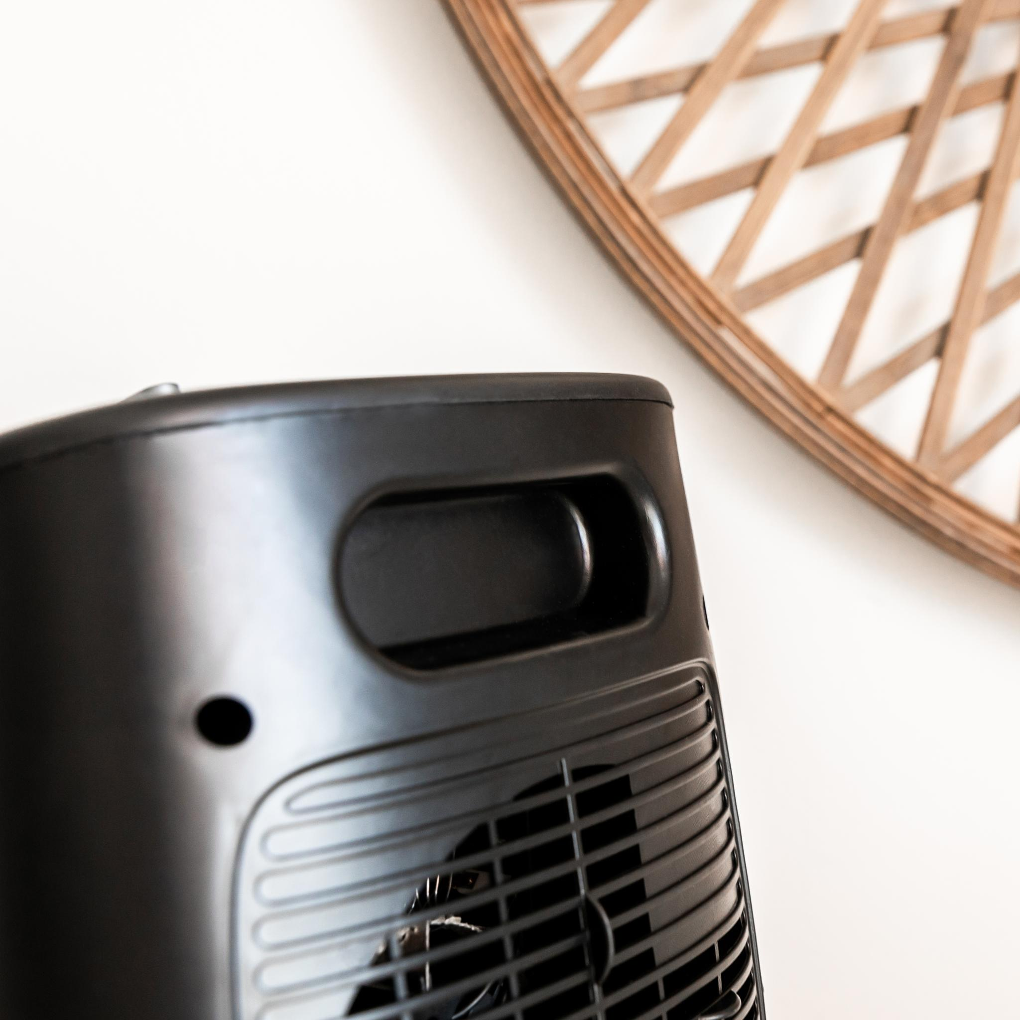 Primer plano de un ventilador negro con asa. Un enrejado de madera decorativo en el fondo borroso contra una pared blanca.