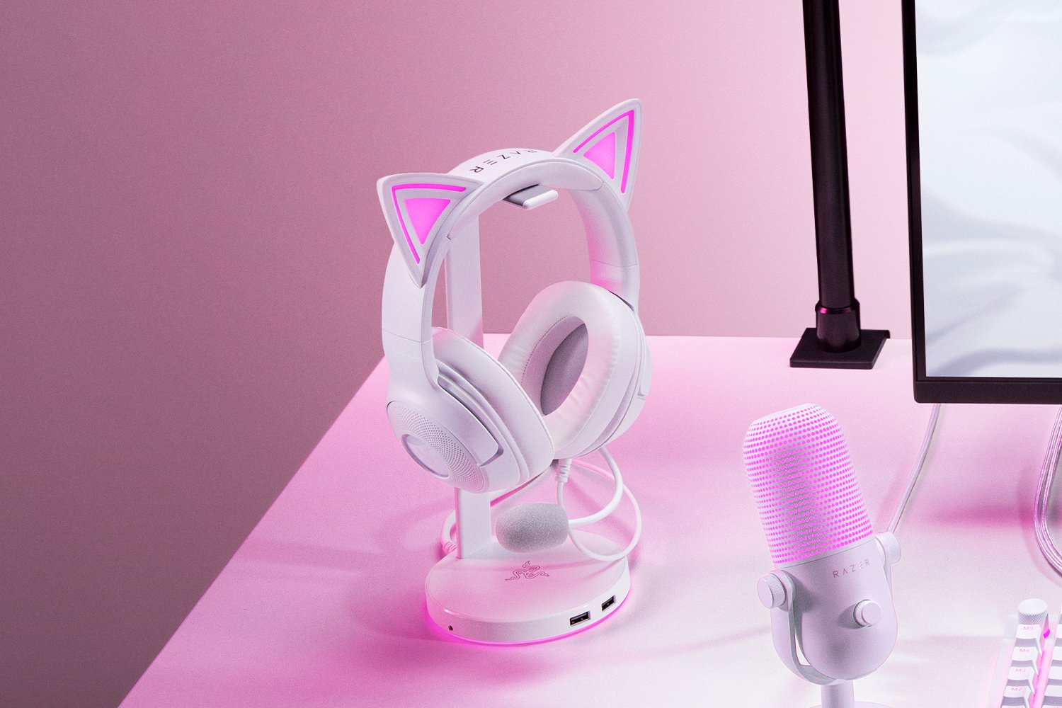 Auriculares blancos con orejas de gato en un soporte, micrófono rosa y un monitor en una mesa rosa. Fondo rosa.