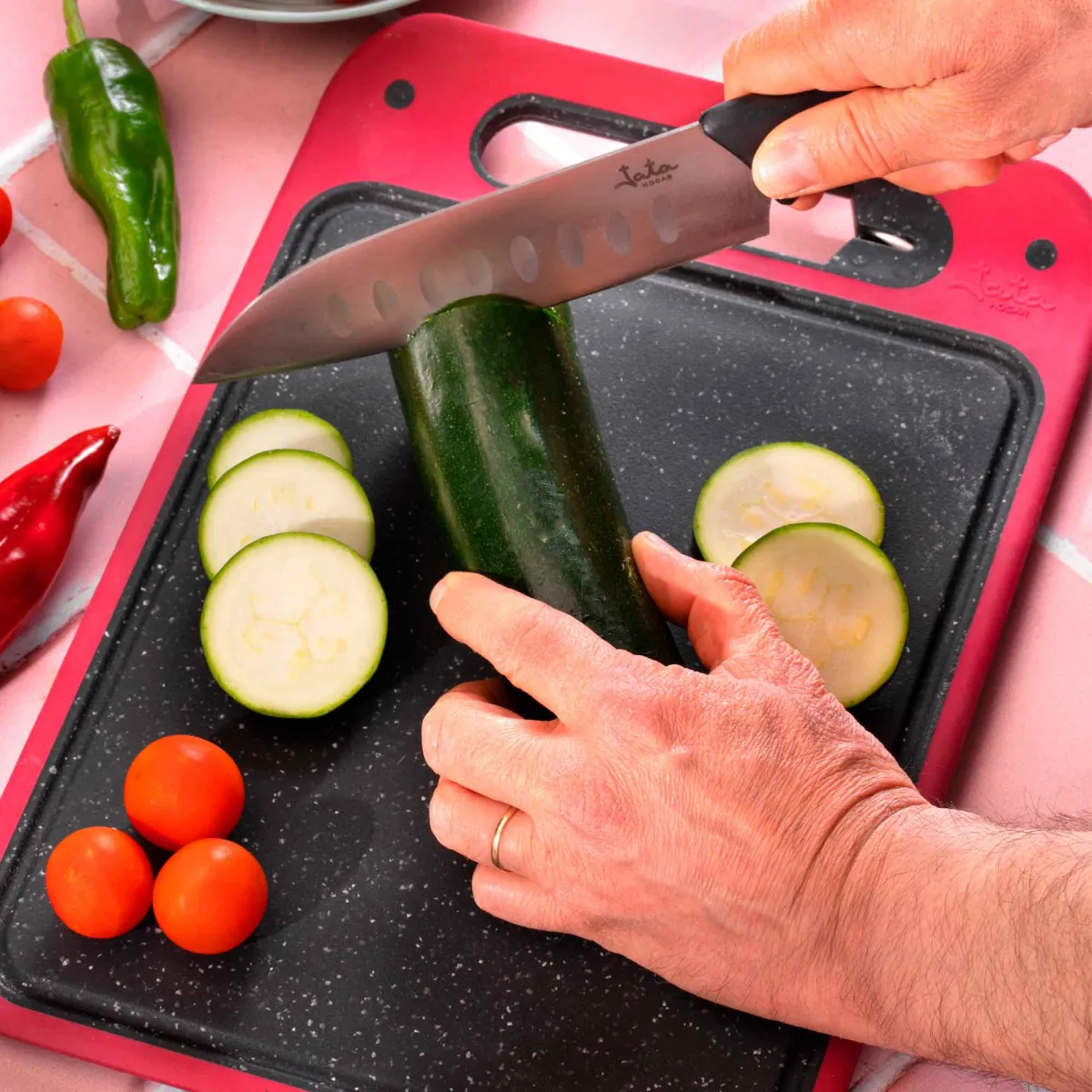 Una persona está cortando un calabacín en una tabla de cortar. Hay calabacín en rodajas, tomates y pimientos.