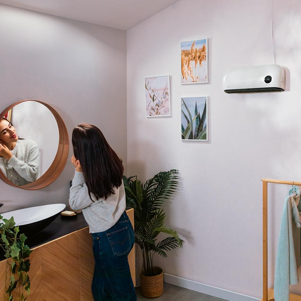 Mujer en un baño mirándose en un espejo redondo. Hay plantas, cuadros y un calefactor en la pared. Lleva vaqueros.