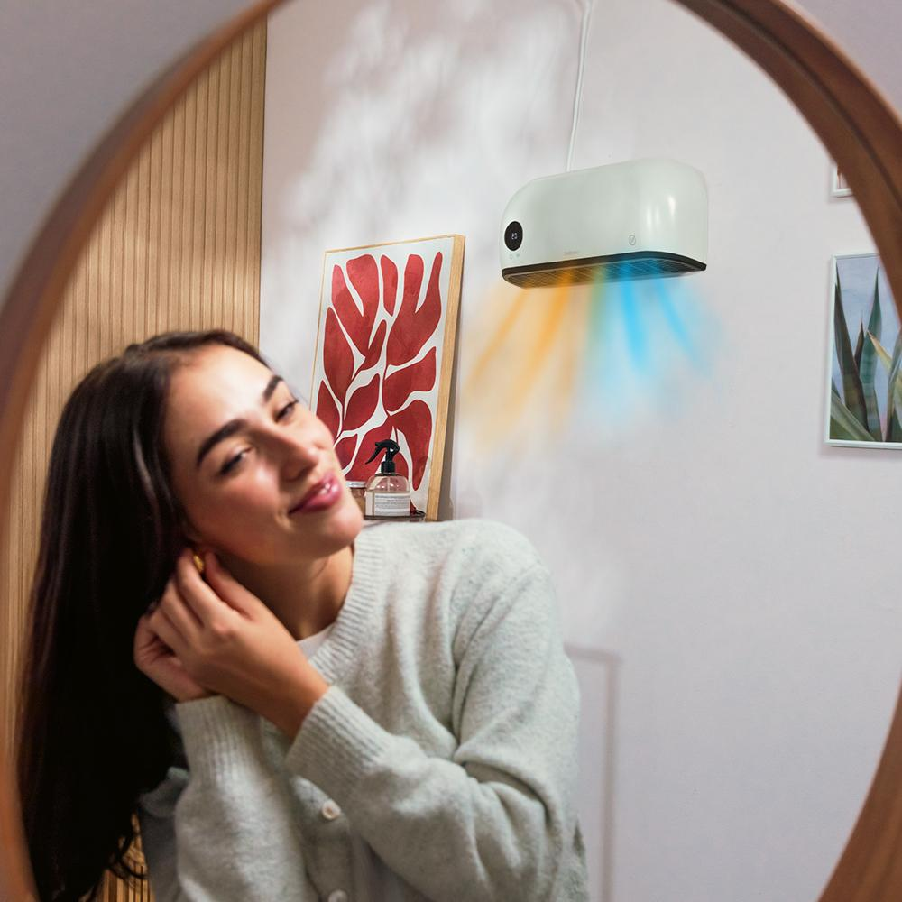 Mujer en espejo poniéndose un pendiente. Purificador de aire blanco en la pared, con corrientes de aire naranja y azul. Obras de arte y decoración en la pared.