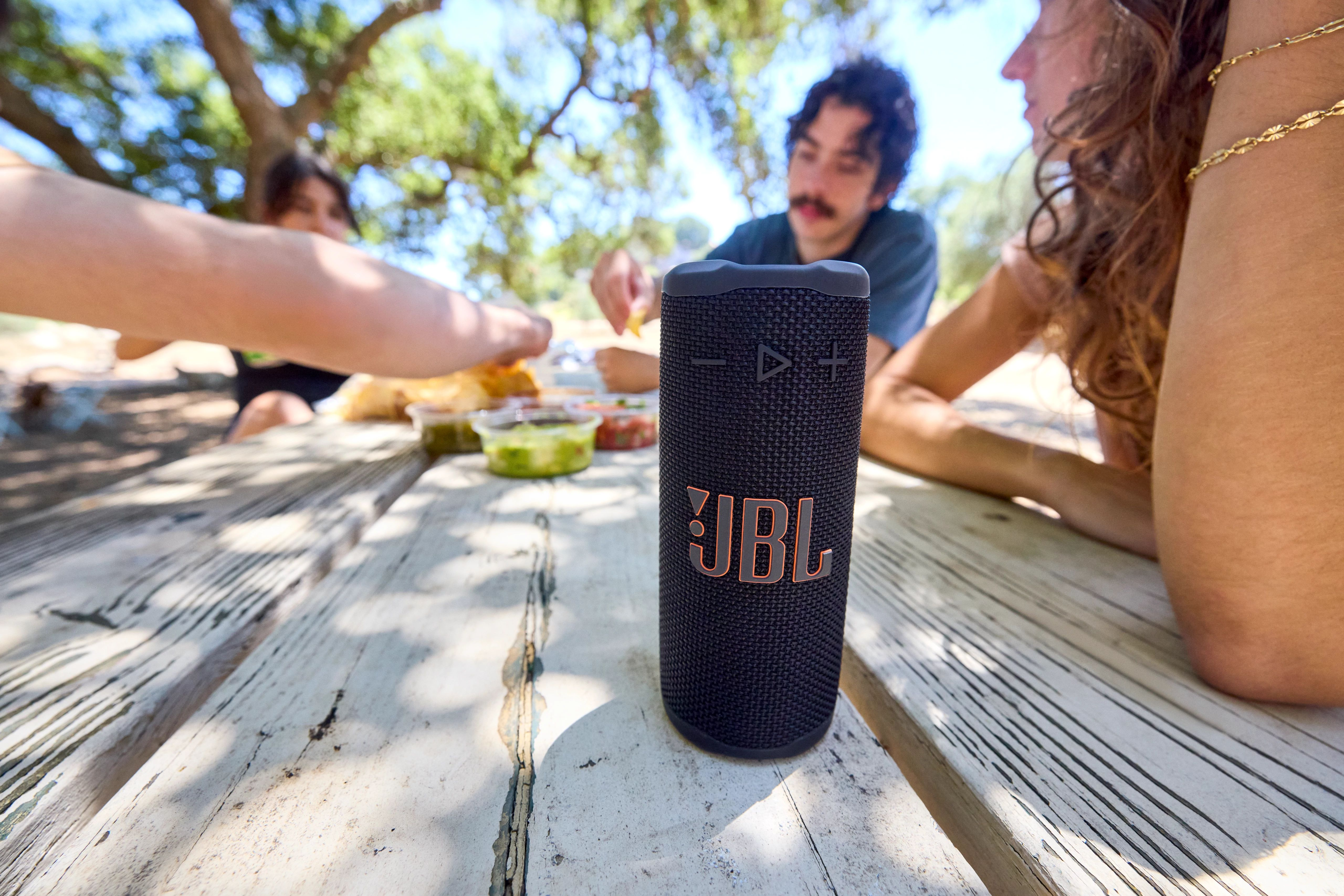 İnsanlar dışarıda yemek yiyor, bir JBL hoparlör masada duruyor. Masada yiyecek ve içecekler var. Arka planda yeşil ağaçlar.