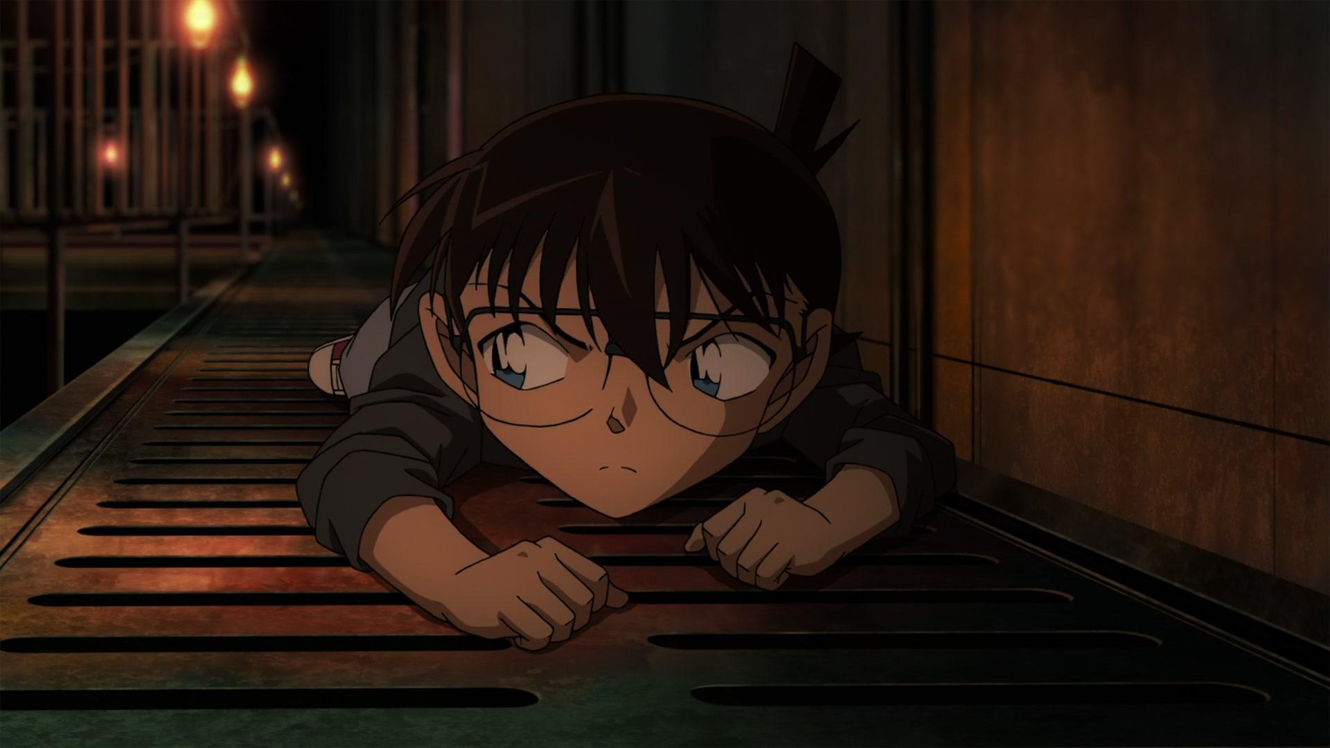 Anime-Figur liegt auf einem Metallgitter und blickt nach unten. Dunkle Haare, Brille und ernster Ausdruck. Schwach beleuchtete Gasse mit Deckenleuchten.