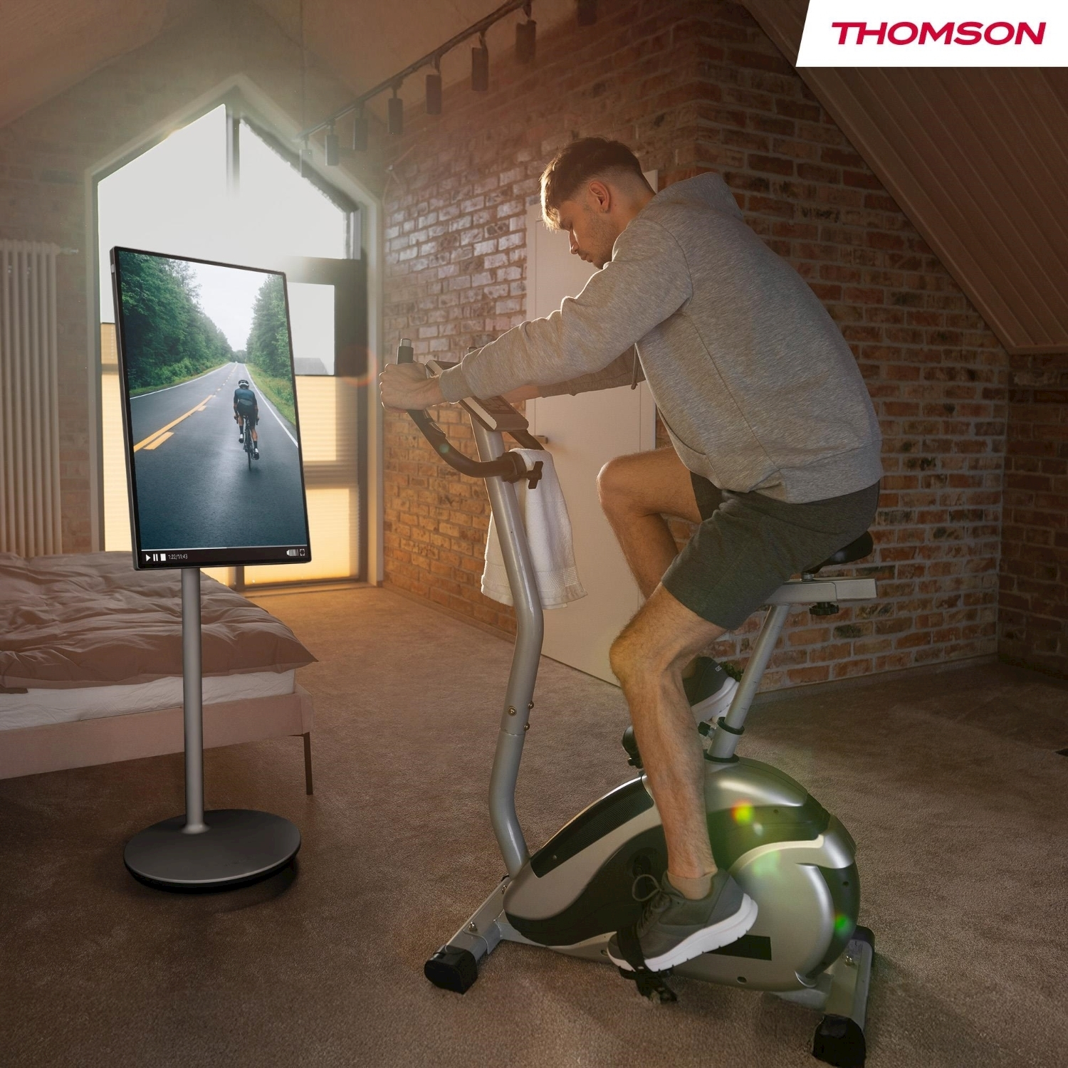 Un uomo si esercita su una cyclette in una stanza. Una TV mostra un ciclista. Muro di mattoni e luce solare presenti.
