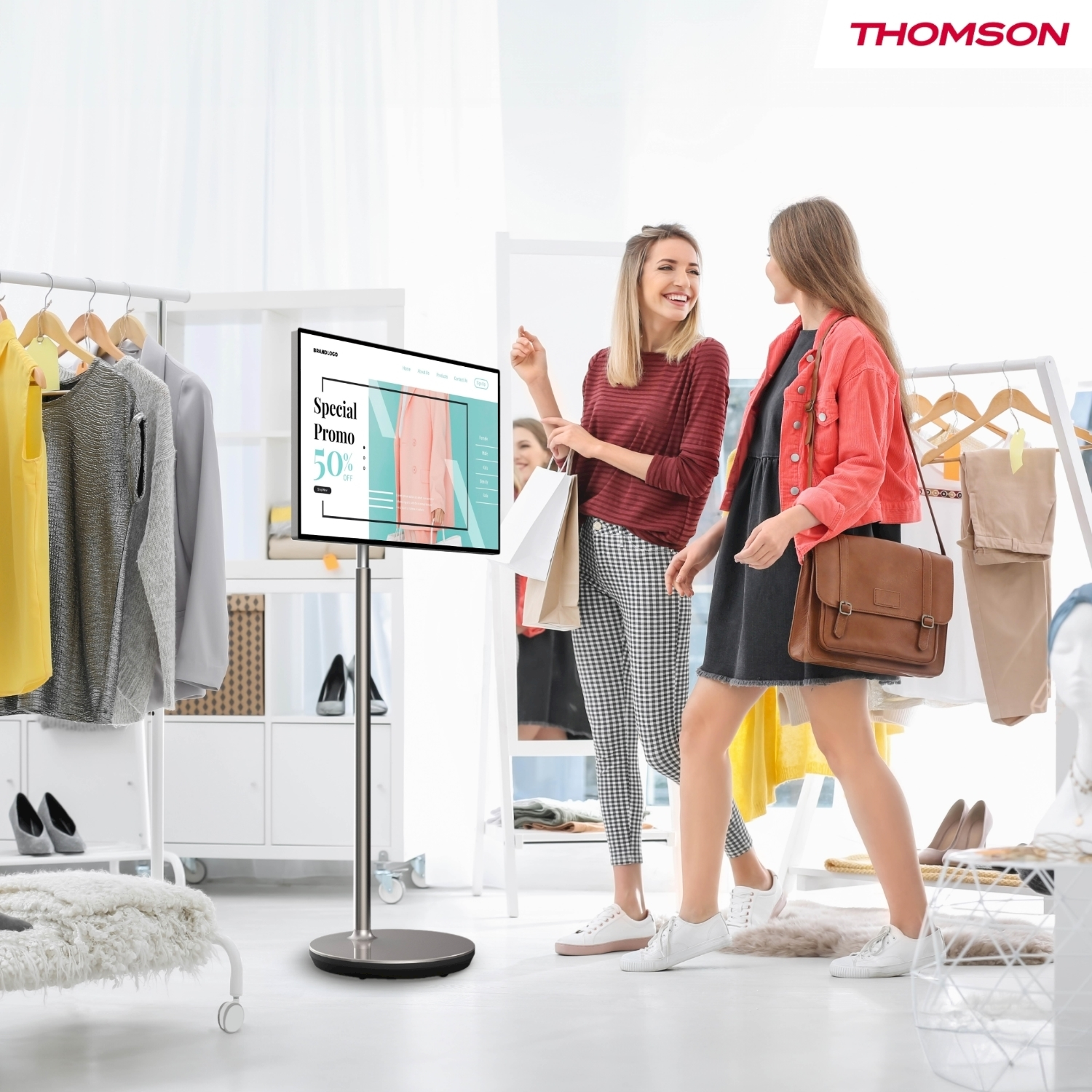 Due donne in un negozio guardano uno schermo che mostra un'offerta. Portabiti e accessori le circondano. Un logo Thomson nell'angolo.