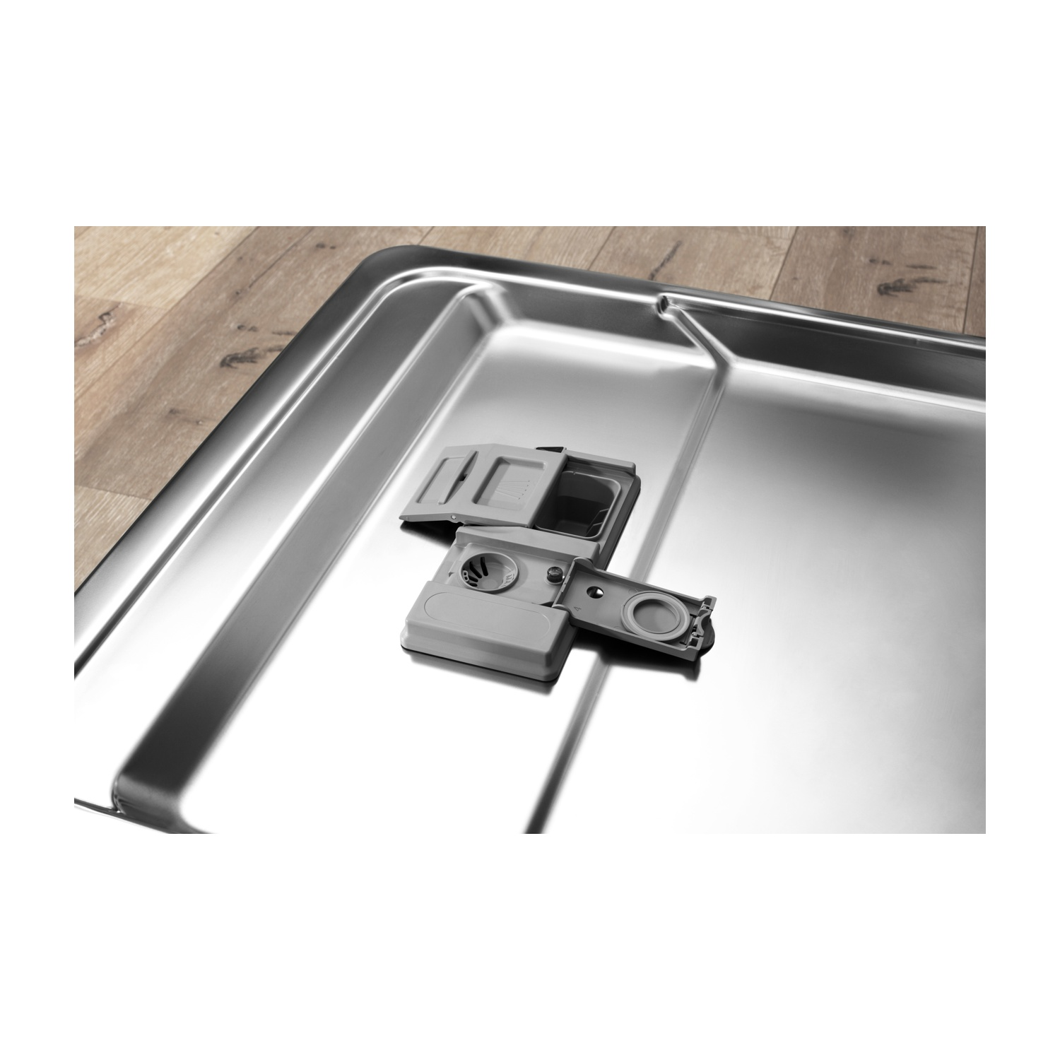 Erogatore di detersivo per lavastoviglie aperto su superficie in acciaio inox. Pavimento in legno visibile sullo sfondo.