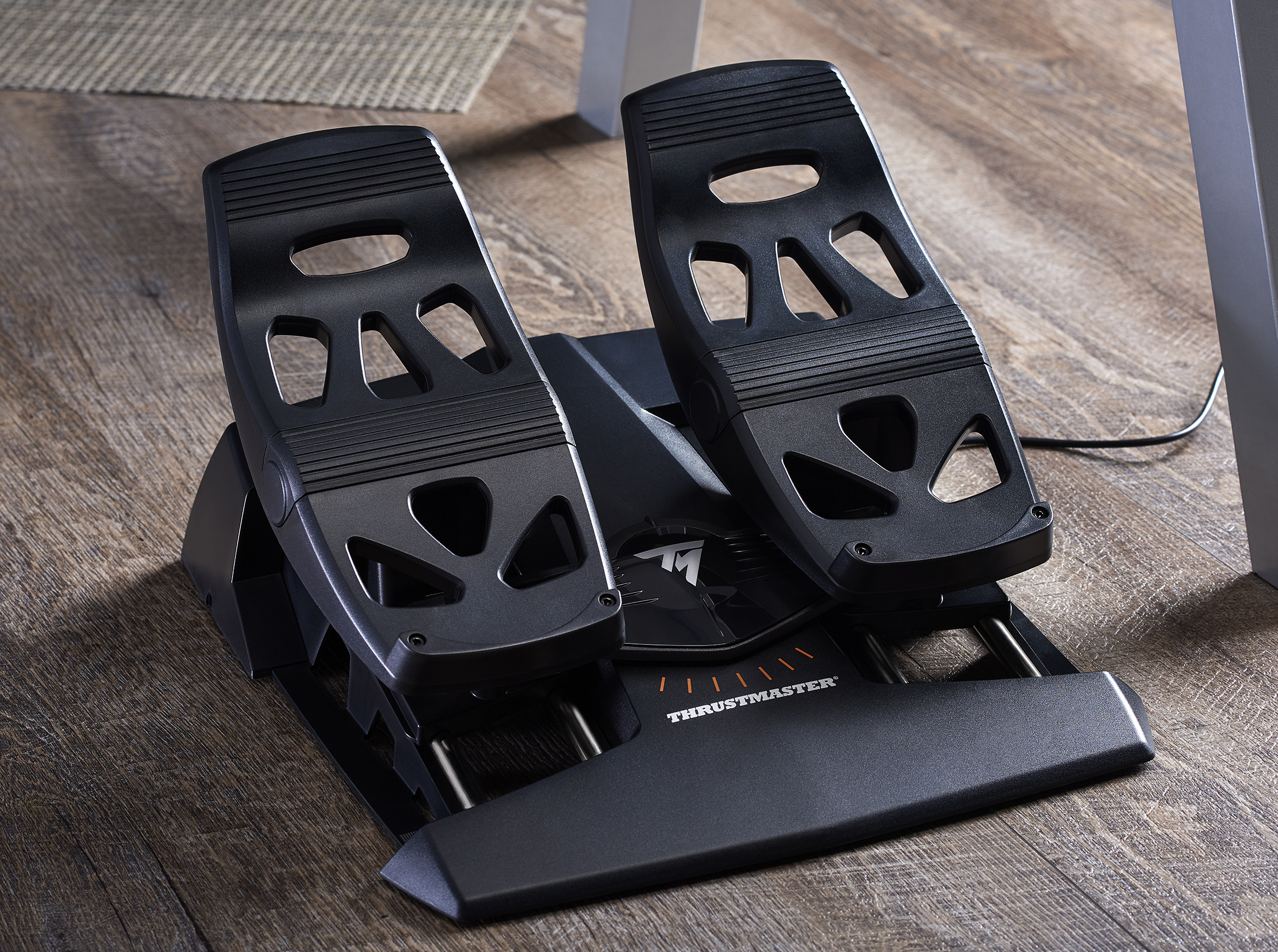 Black Thrustmaster racing pedals on a wooden floor, near a white table leg. The pedals are black and detailed.